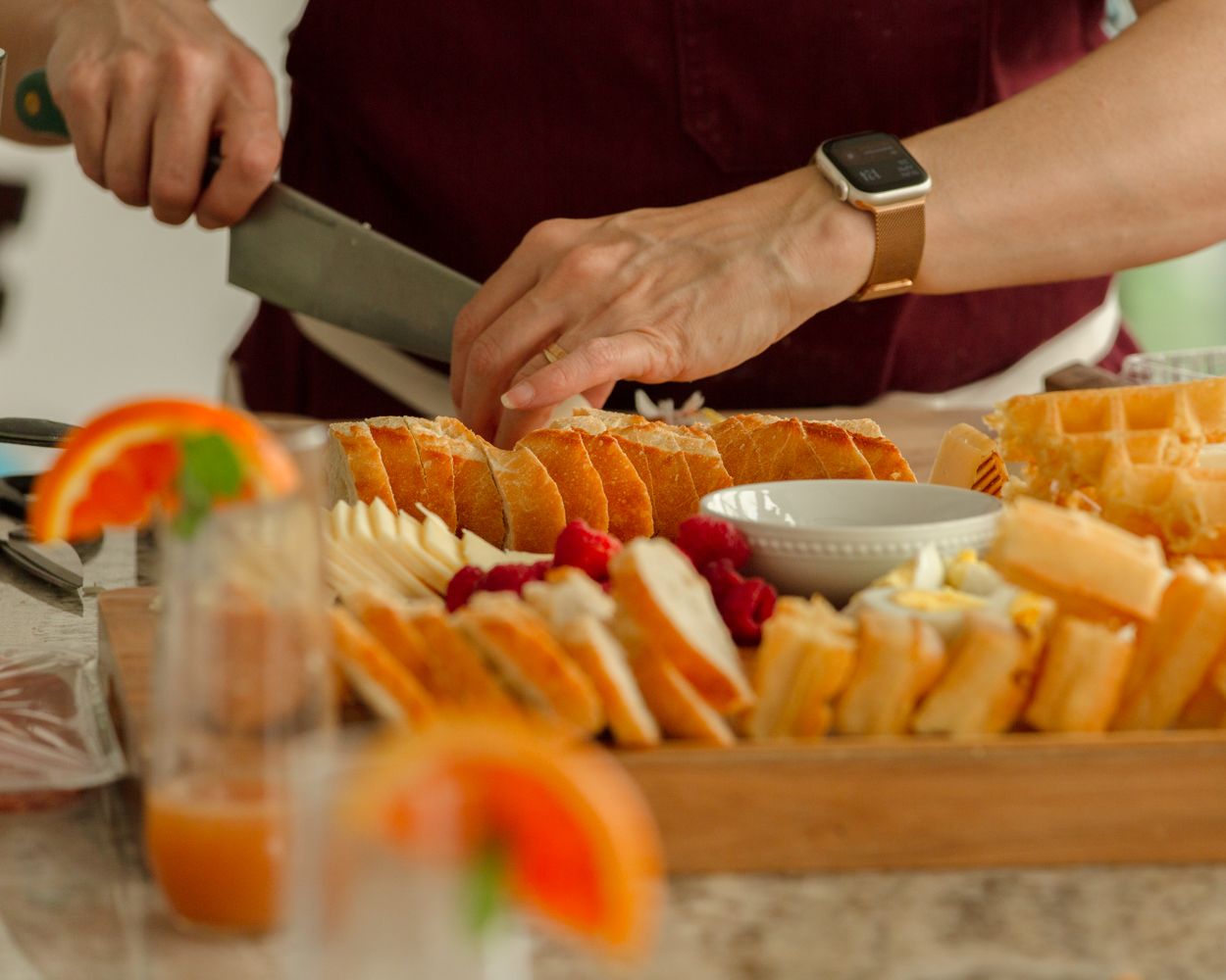 Person arranging a charcuterie board with waffles, cheese, strawberries, and grapefruit slices on a wooden tray.