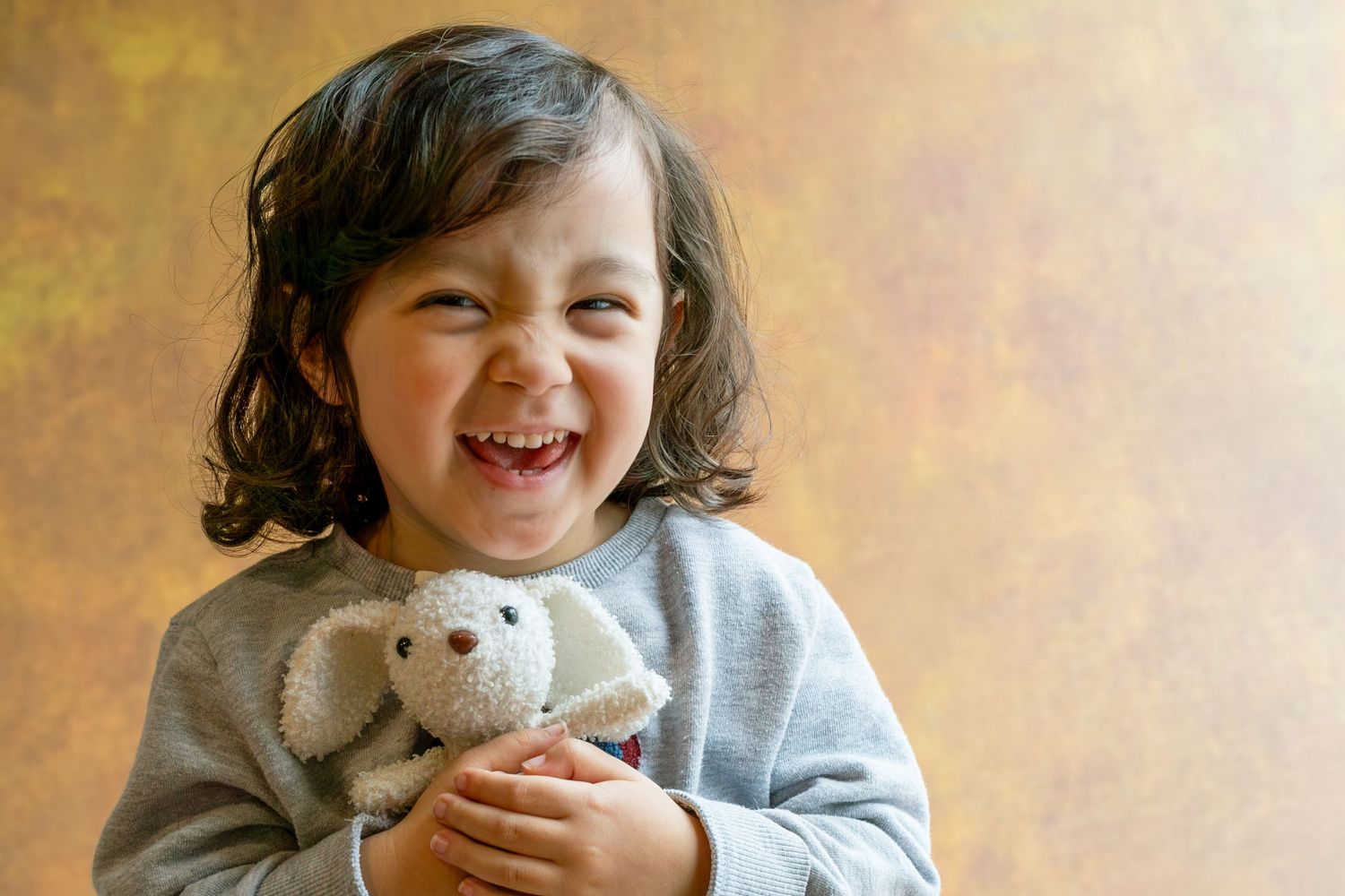 A joyful child wearing a gray sweater smiles brightly while holding a small stuffed animal toy.