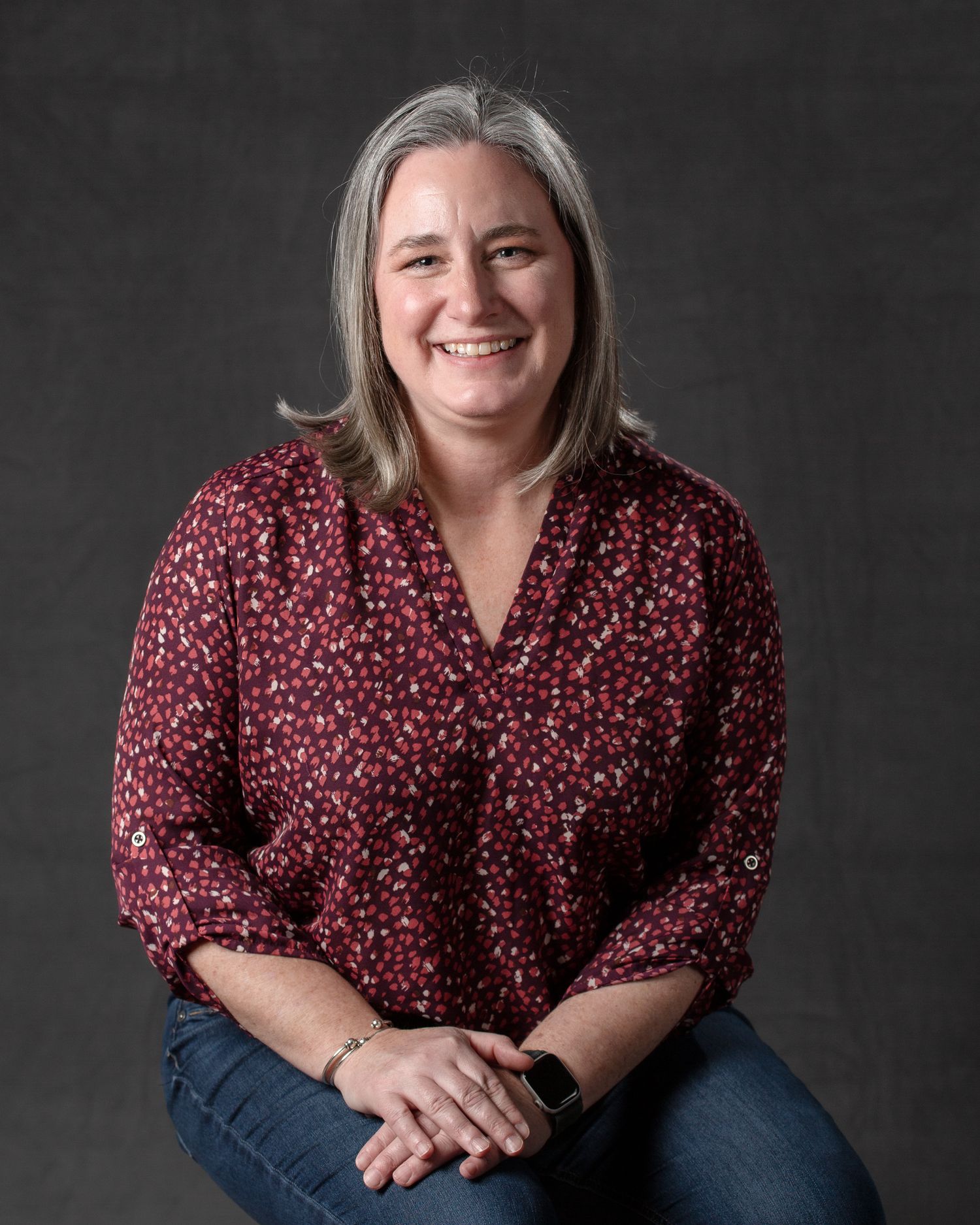 Smiling woman with gray-streaked hair wearing a red floral blouse and jeans, seated against a gray studio background.