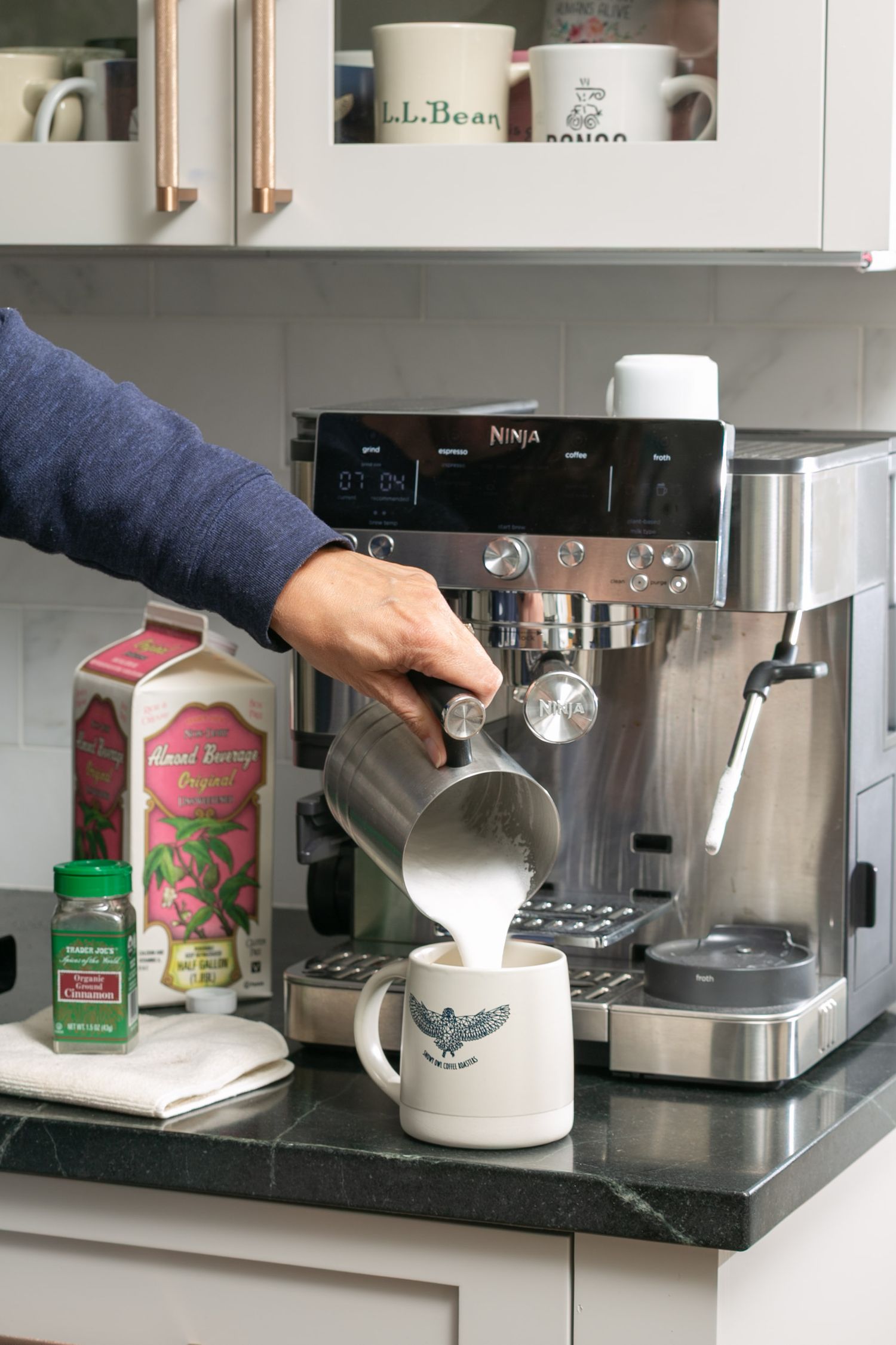 Person pouring steamed milk into a mug using a Ninja espresso machine on a kitchen counter.