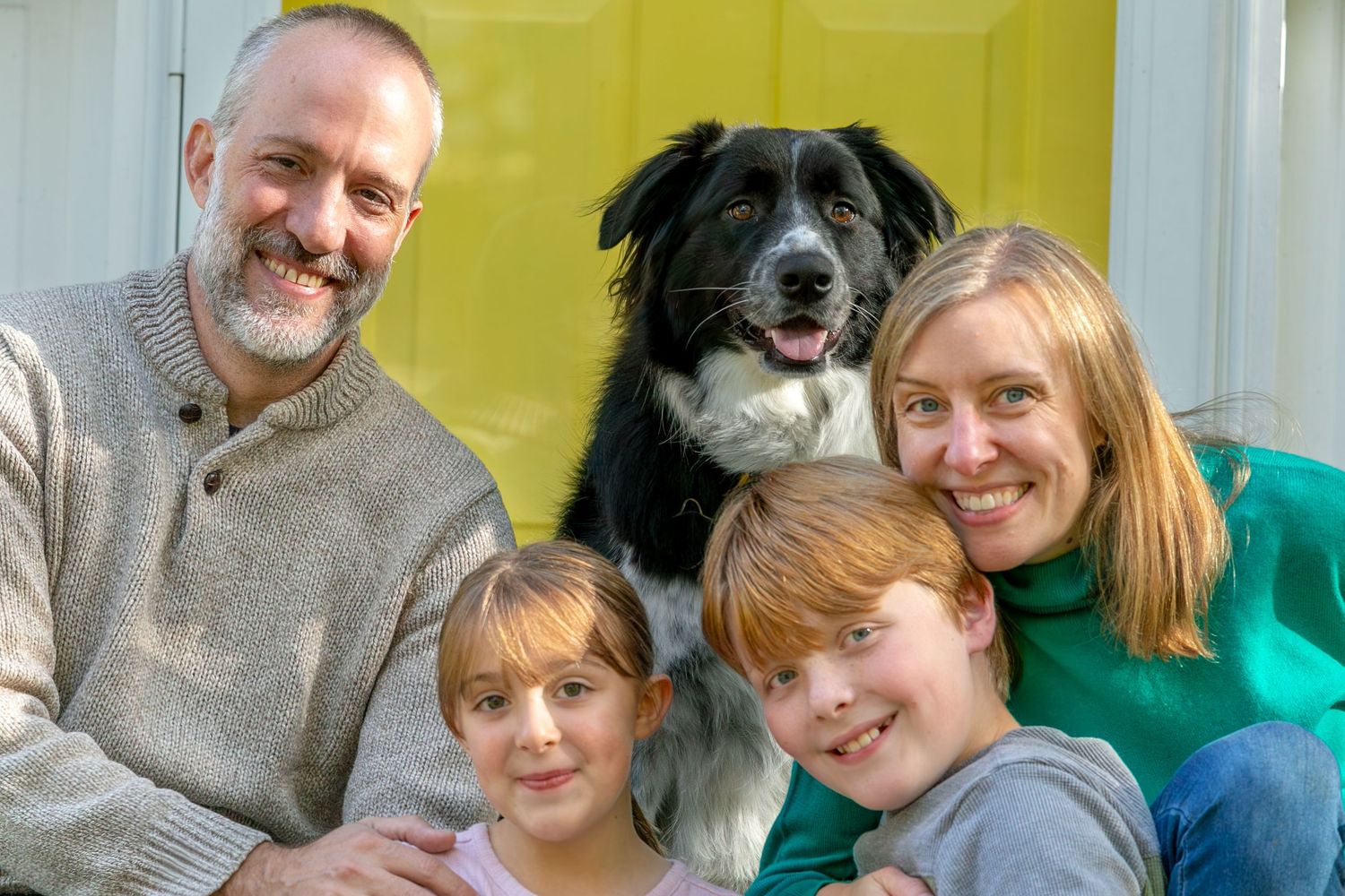 A happy family poses together with their black and white Border Collie dog against a yellow background.