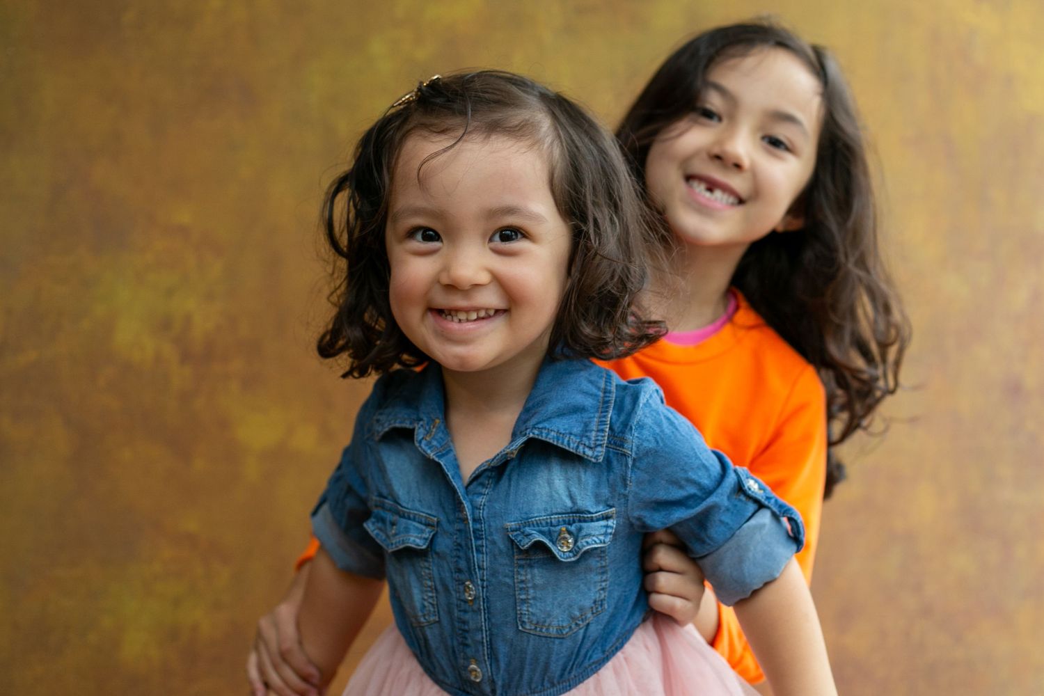 Two children smile and laugh together while wearing casual clothing against a warm brown backdrop.