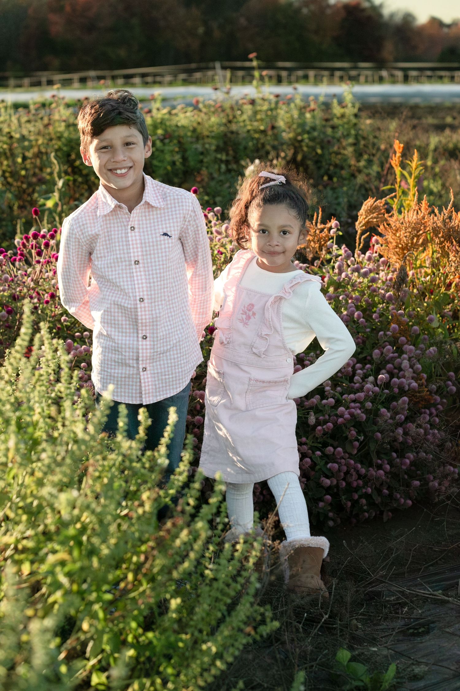 Two children stand together in a garden with blooming flowers and golden sunlight during sunset.