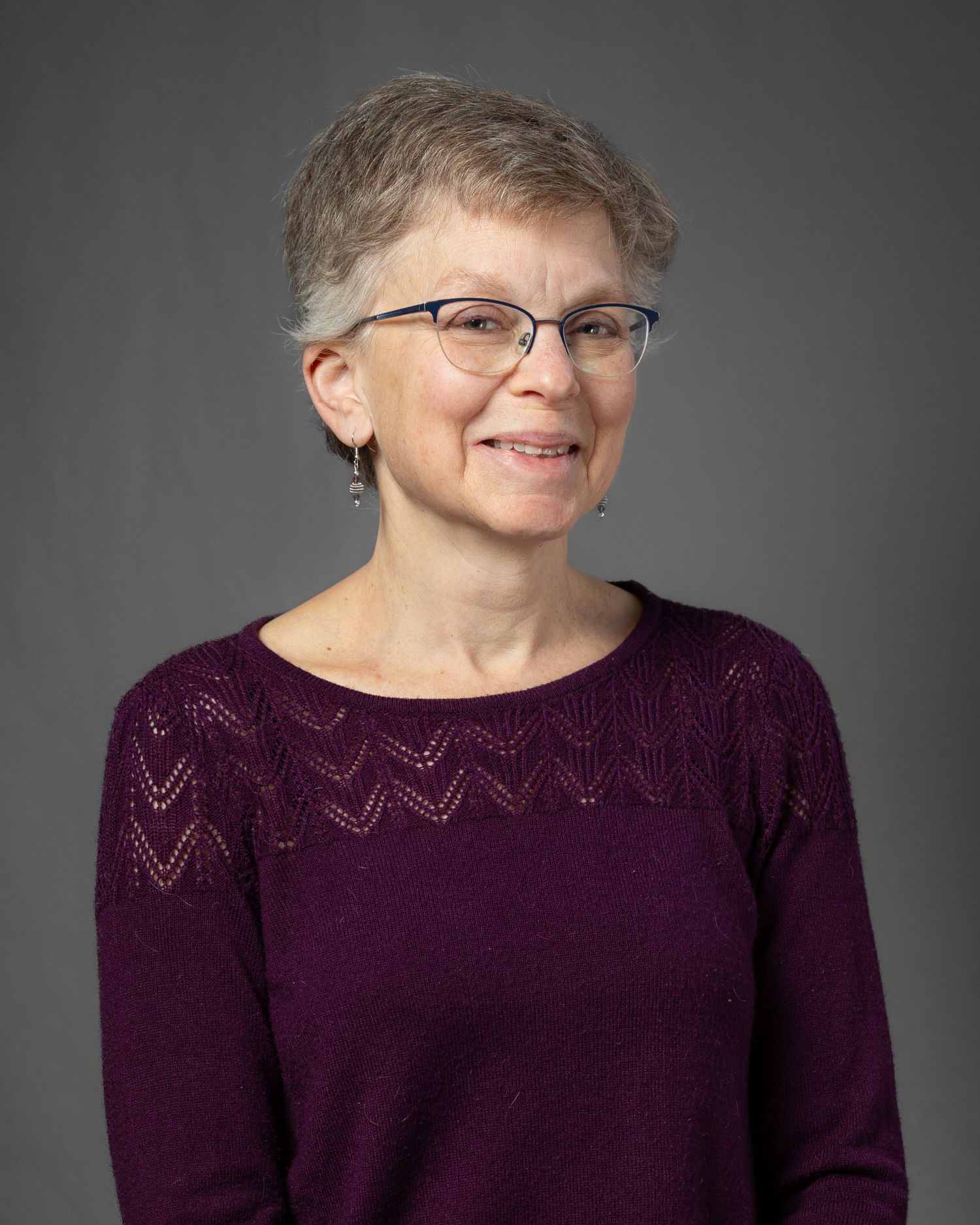 A person wearing glasses and a purple sweater smiles warmly against a gray studio backdrop.