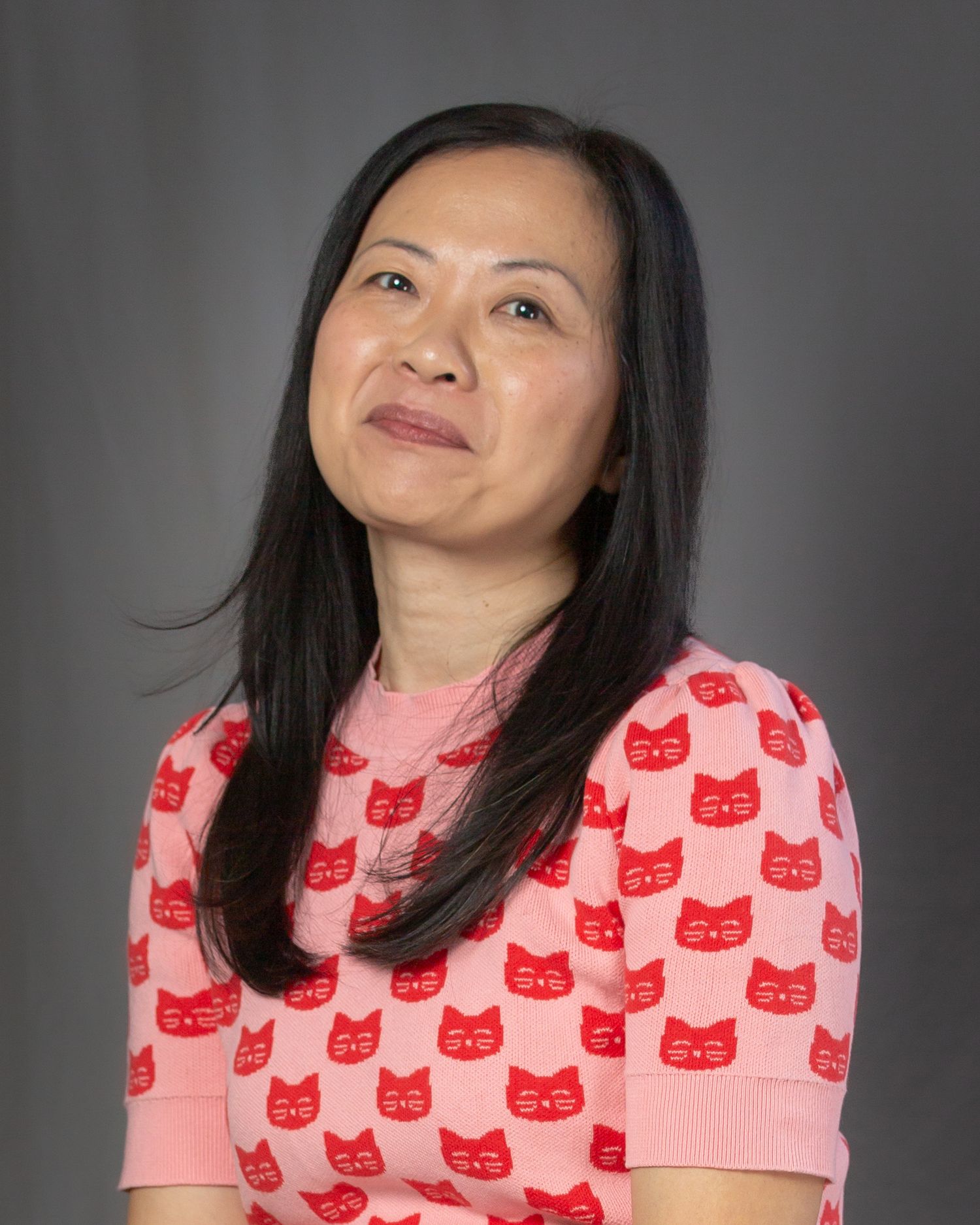 Person wearing pink shirt with repeating red cat pattern makes playful expressions against gray background.