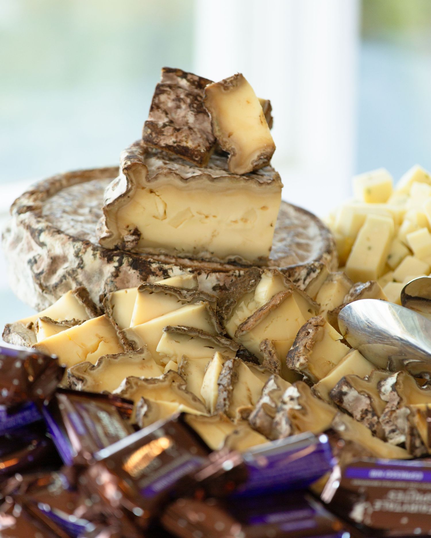 Stacked artisan cheese wheel with rustic rind, surrounded by cut cheese pieces and chocolate candies on a serving platter.