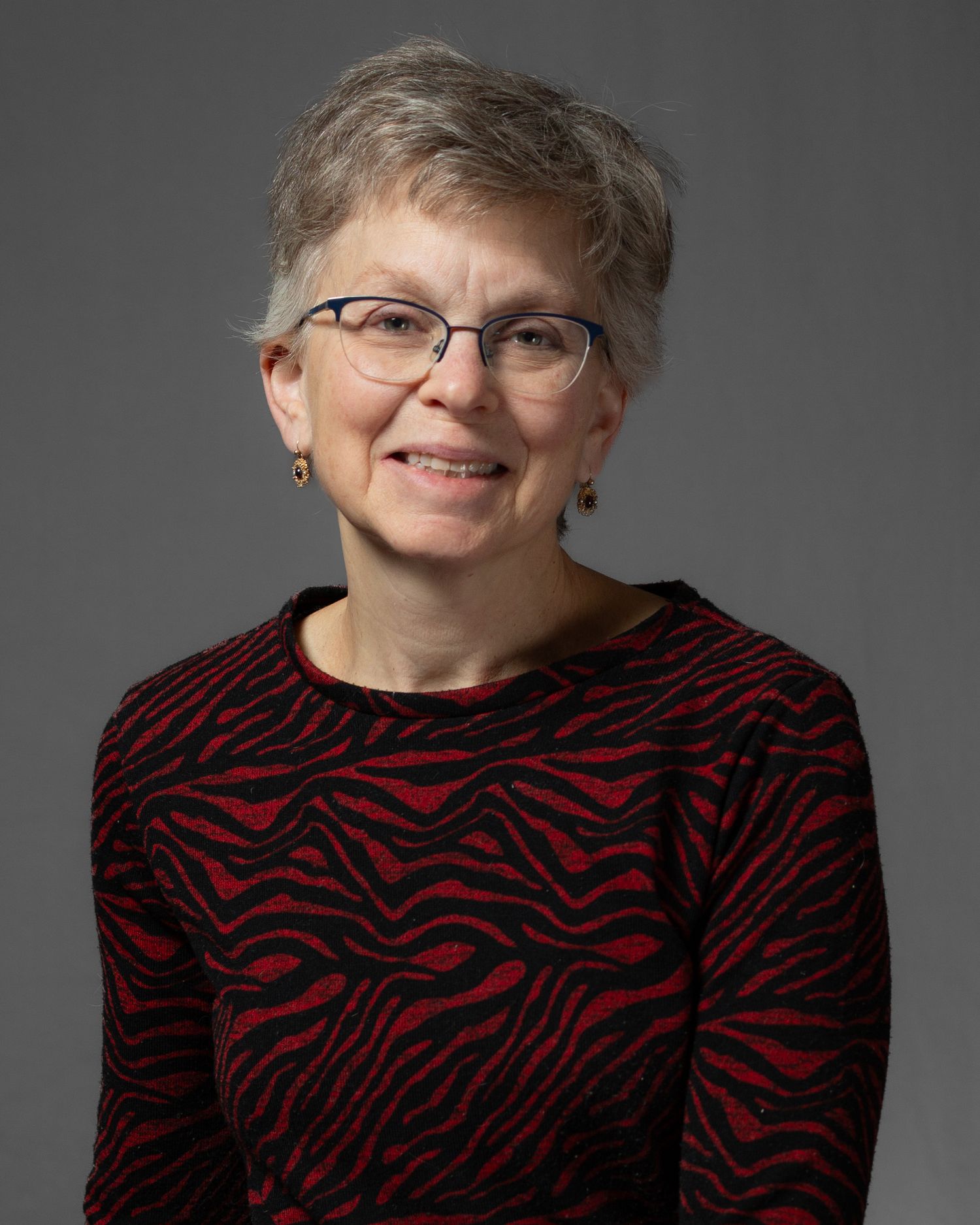 A person wearing glasses and a red and black patterned sweater poses for a series of headshots against a gray background.