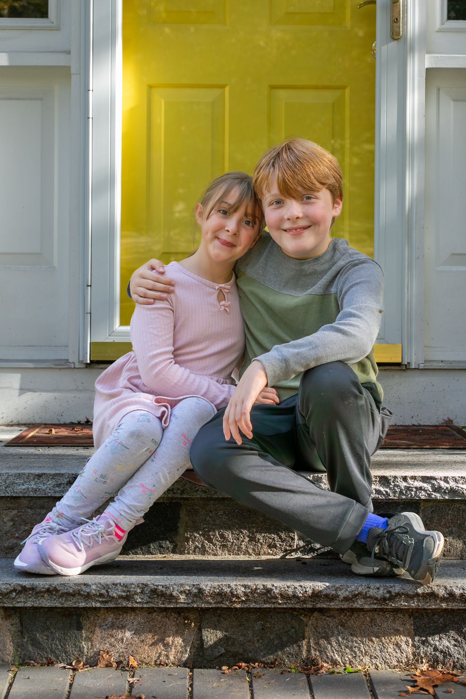 Two young children sit together on a front porch step wearing casual clothing on an autumn day.