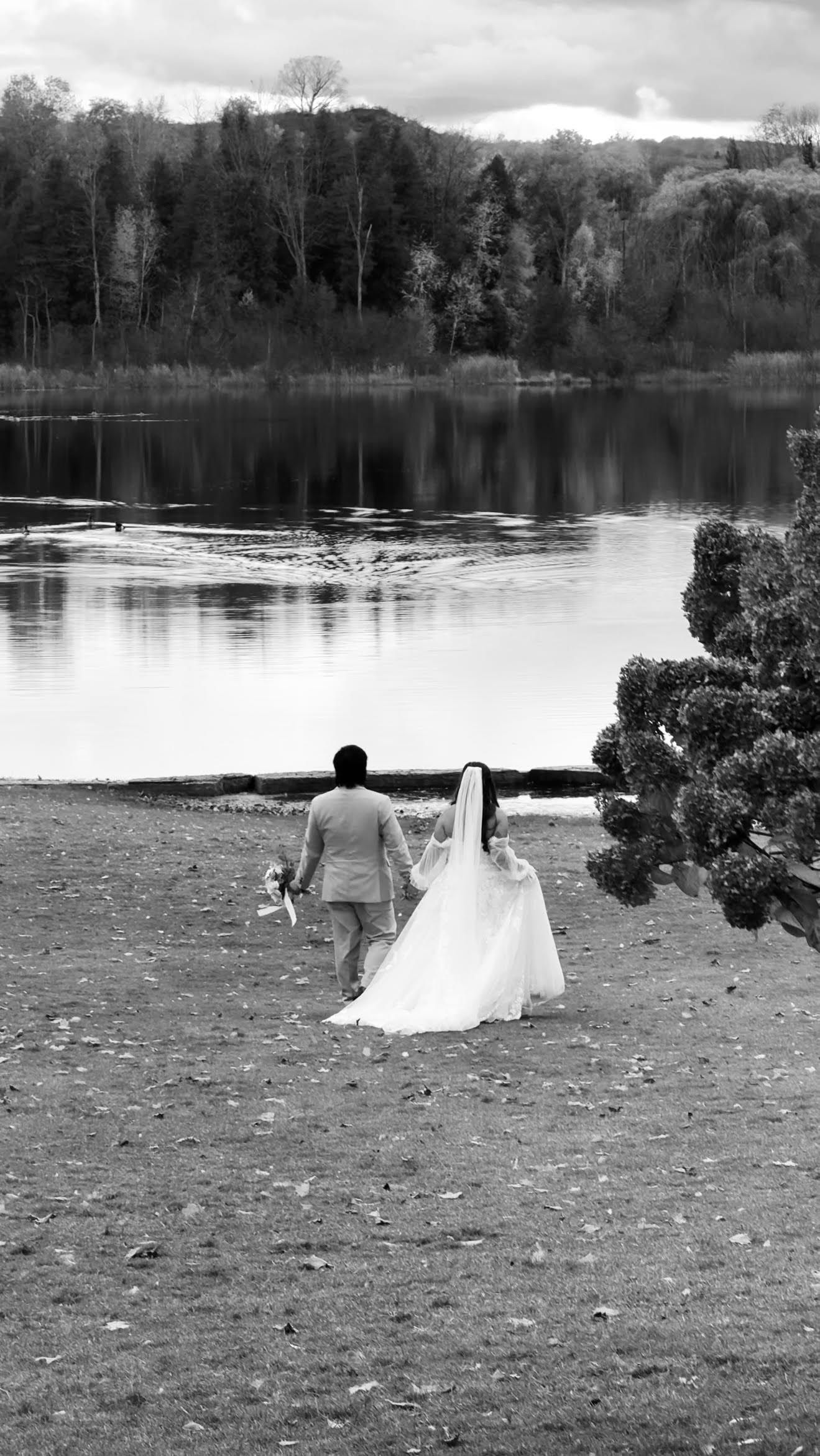 A black and white wedding photo captures a couple lying on grass near a peaceful lake.