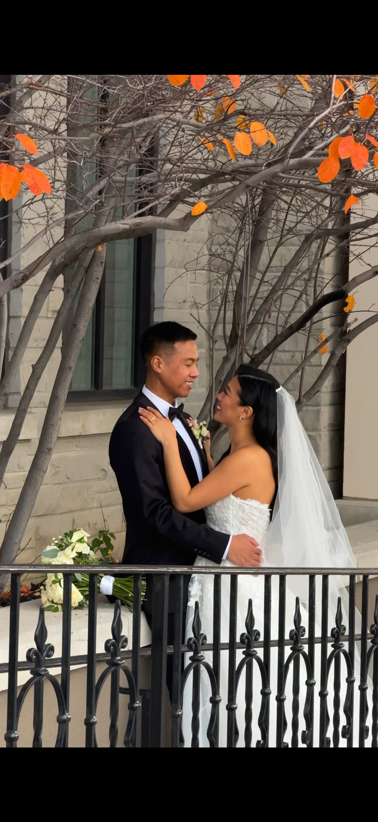 Newlyweds share an intimate moment on a terrace with autumn leaves and an iron railing in the background.