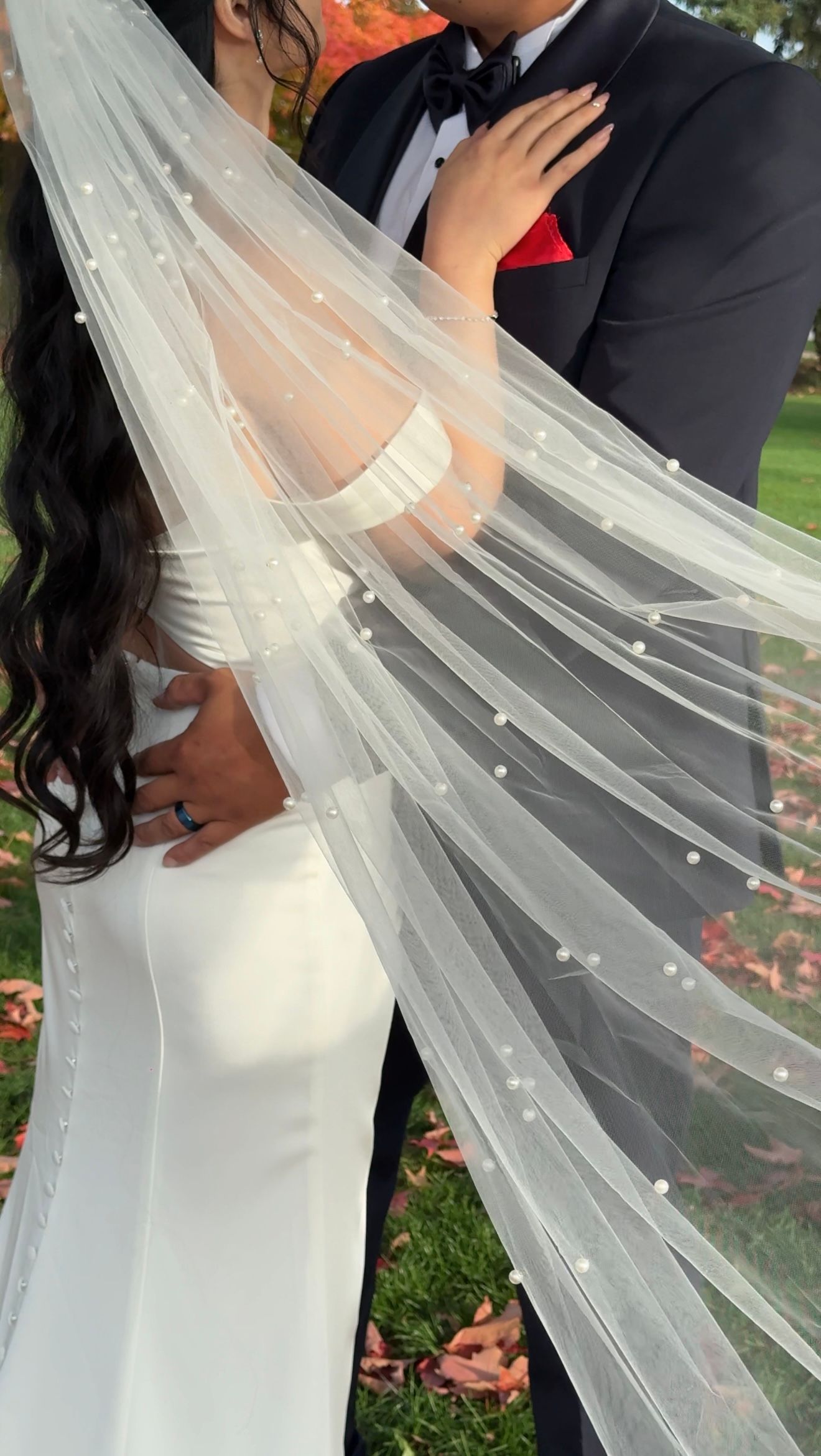 A close-up wedding photo showing a flowing pearl-studded veil and white bridal gown against a black tuxedo with red pocket square.