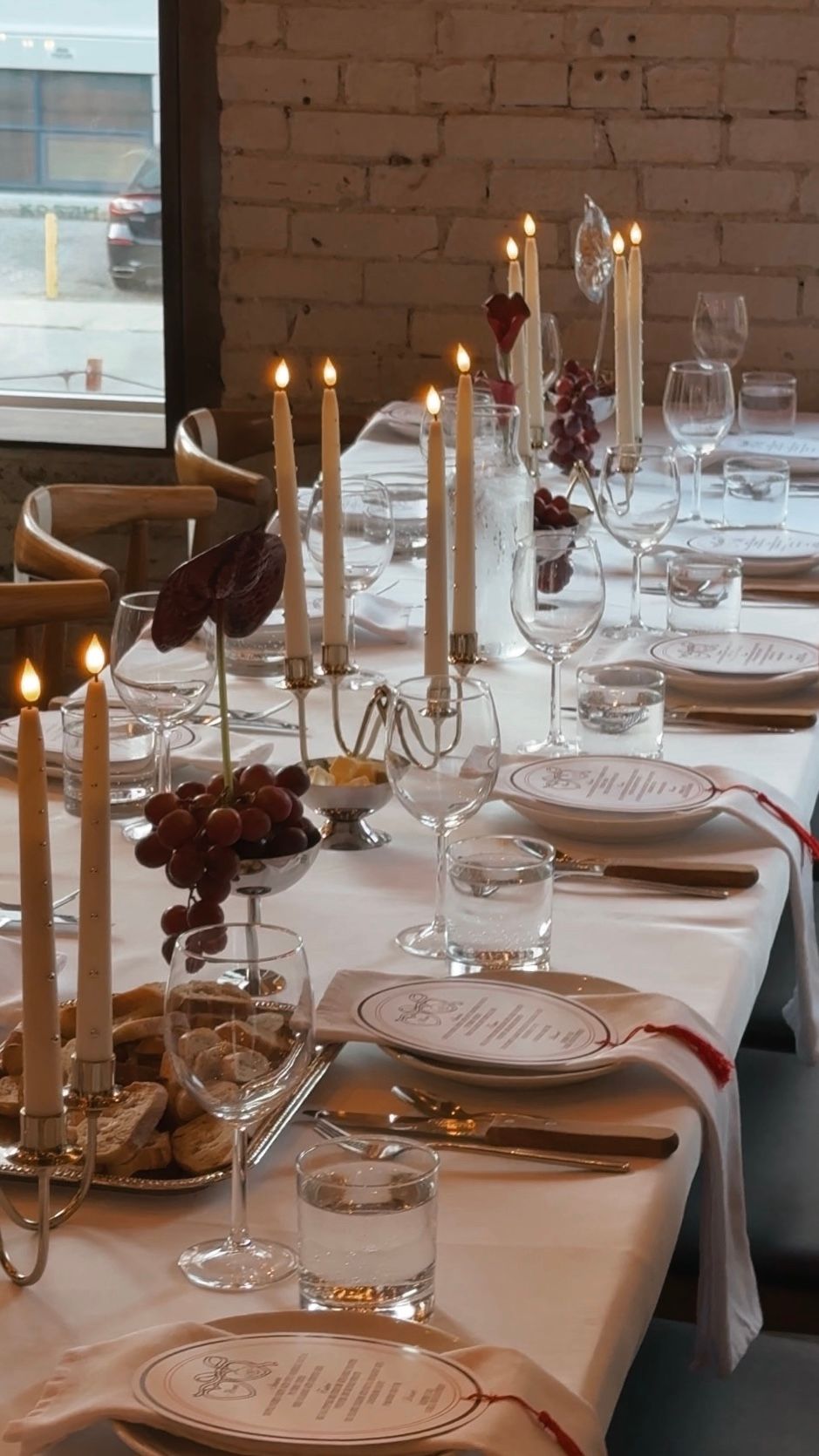 Elegant table setting with white linens, candles, wine glasses and place settings in a rustic brick-walled dining space.