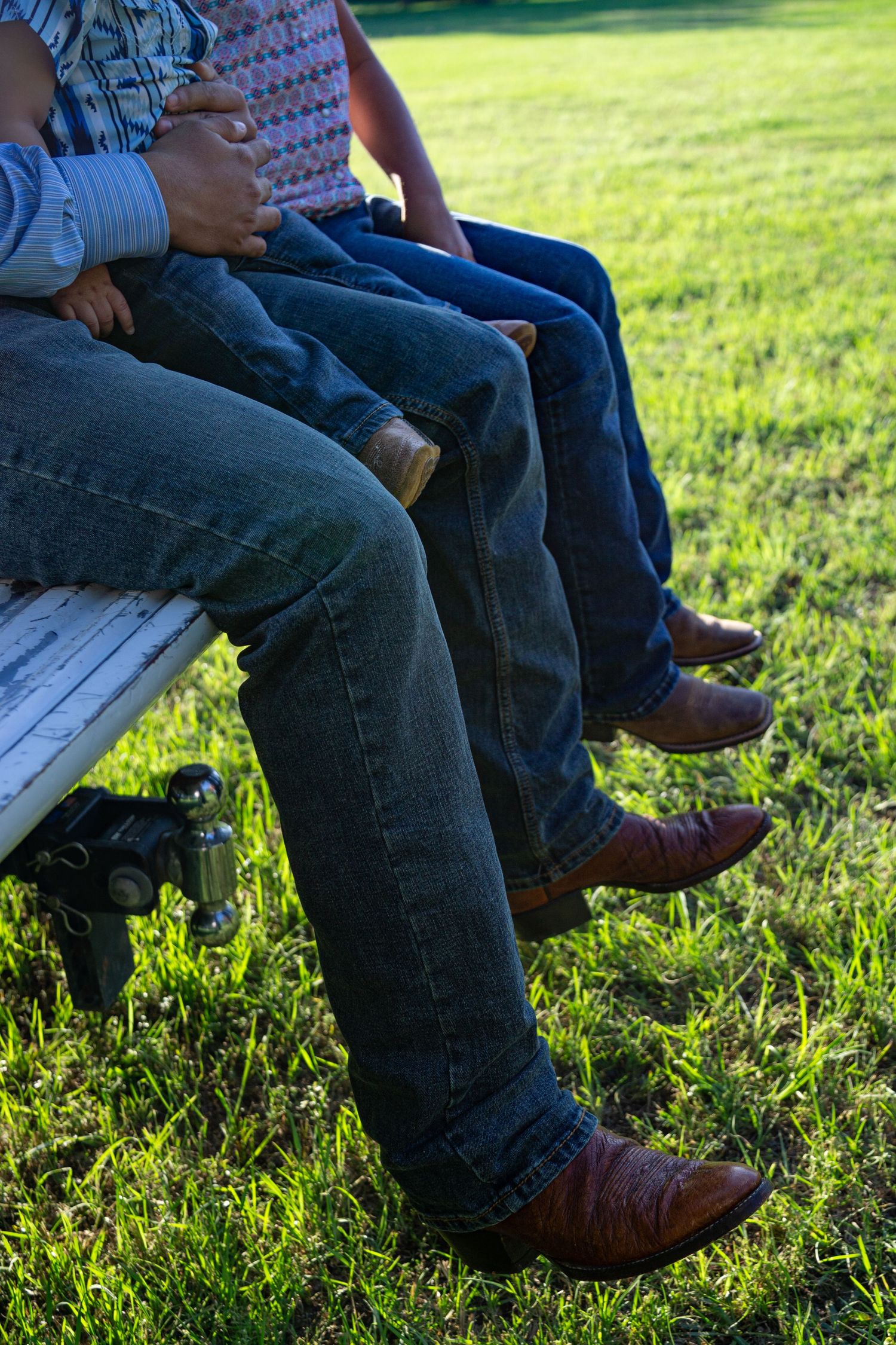Two people in jeans and brown cowboy boots sitting on a bench outdoors on green grass.