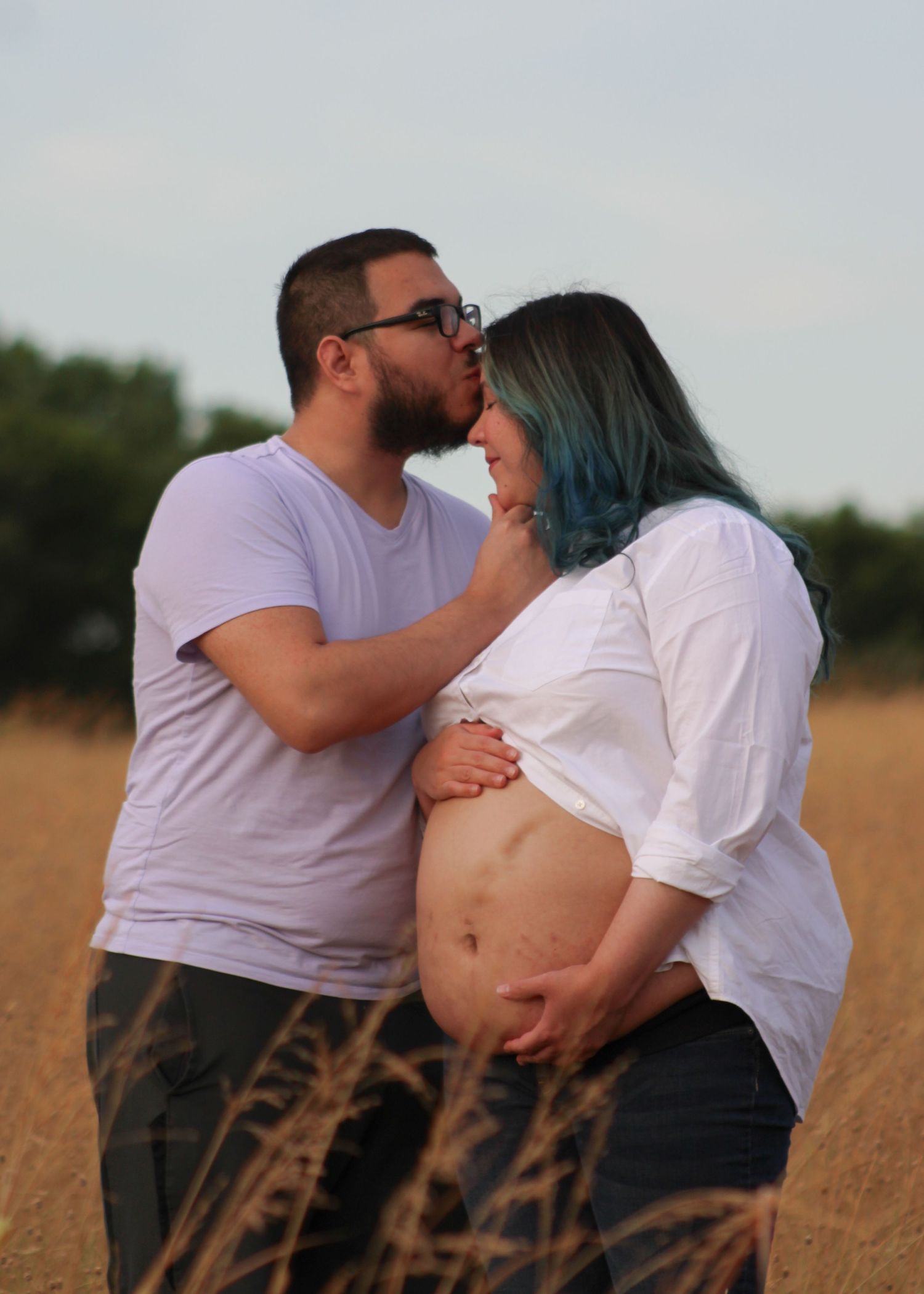 A couple in white shirts share intimate moments in a golden wheat field during a maternity photo session.