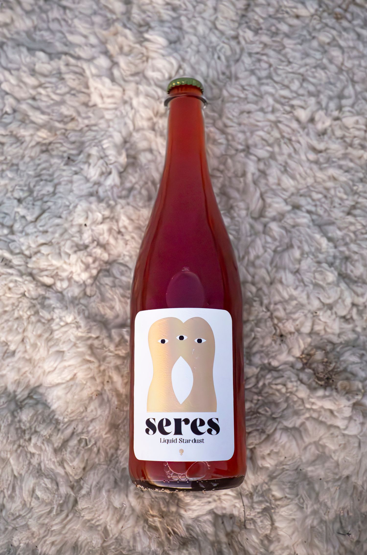 Seres Liquid Stardust natural wine in a pink-red bottle with whimsical character label on textured surface.