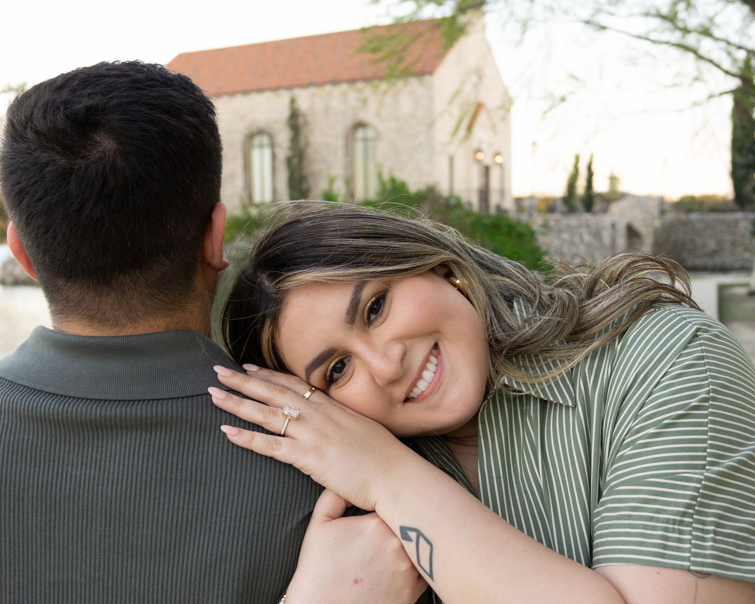 Smiling woman rests her head on man's shoulder outdoors, showing engagement ring near stone building.