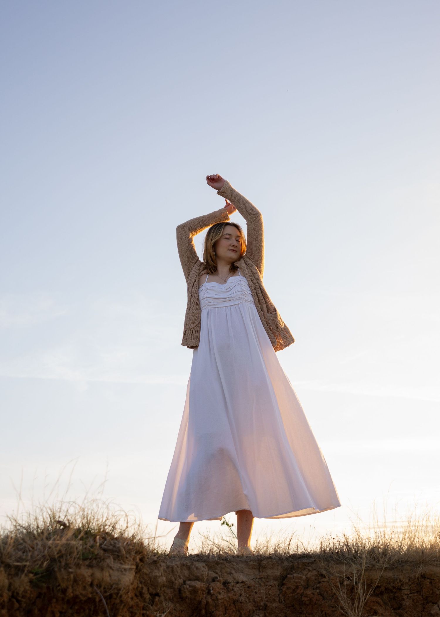 Figure in a flowing white maxi dress and brown cardigan poses gracefully against sunset sky on hilltop.