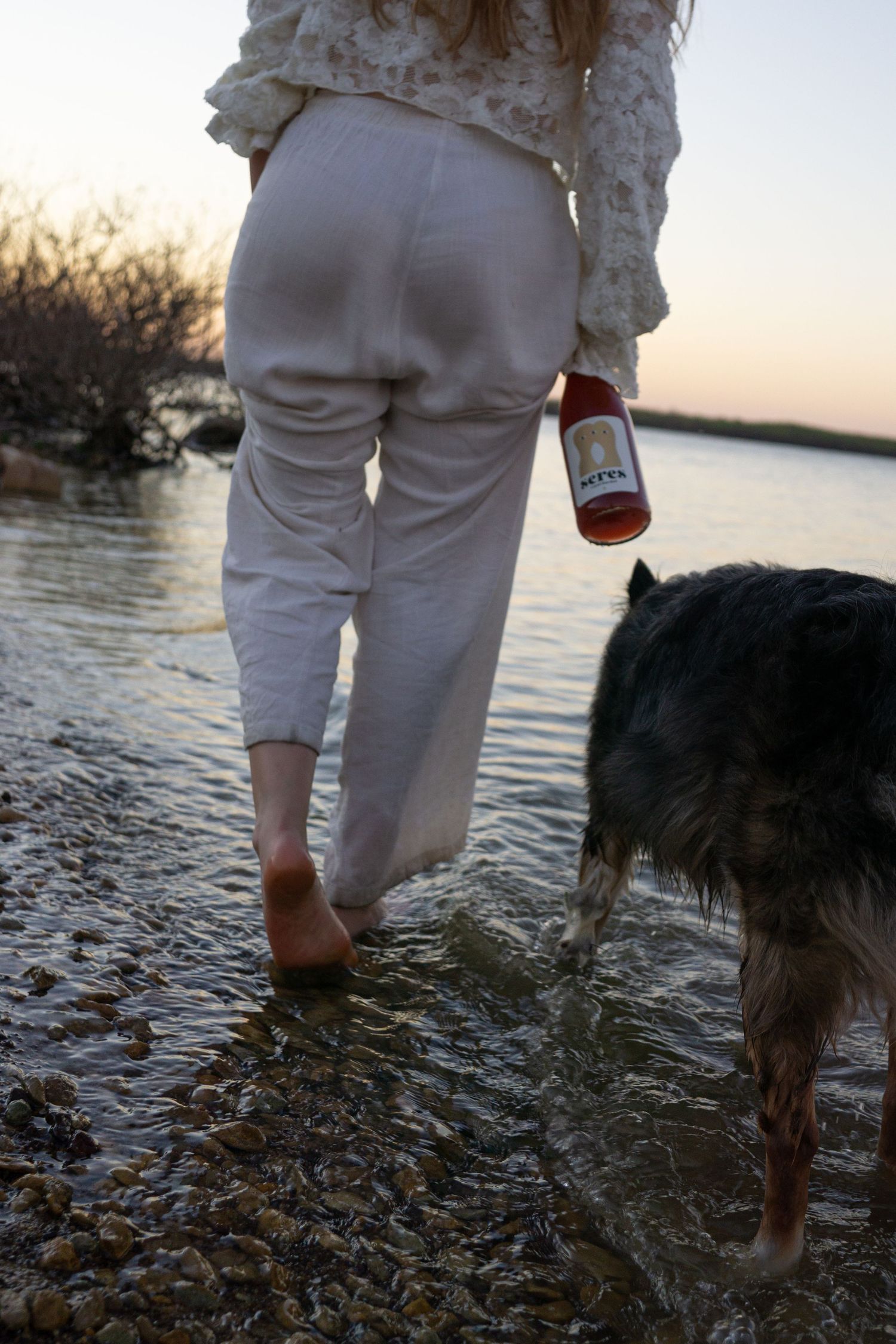 Woman in white linen pants wading barefoot in shallow water at sunset, holding a bottle, with a dog beside her.