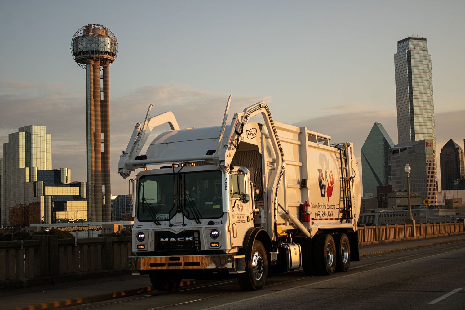 White garbage truck driving on bridge with Dallas skyline and Reunion Tower in background.
