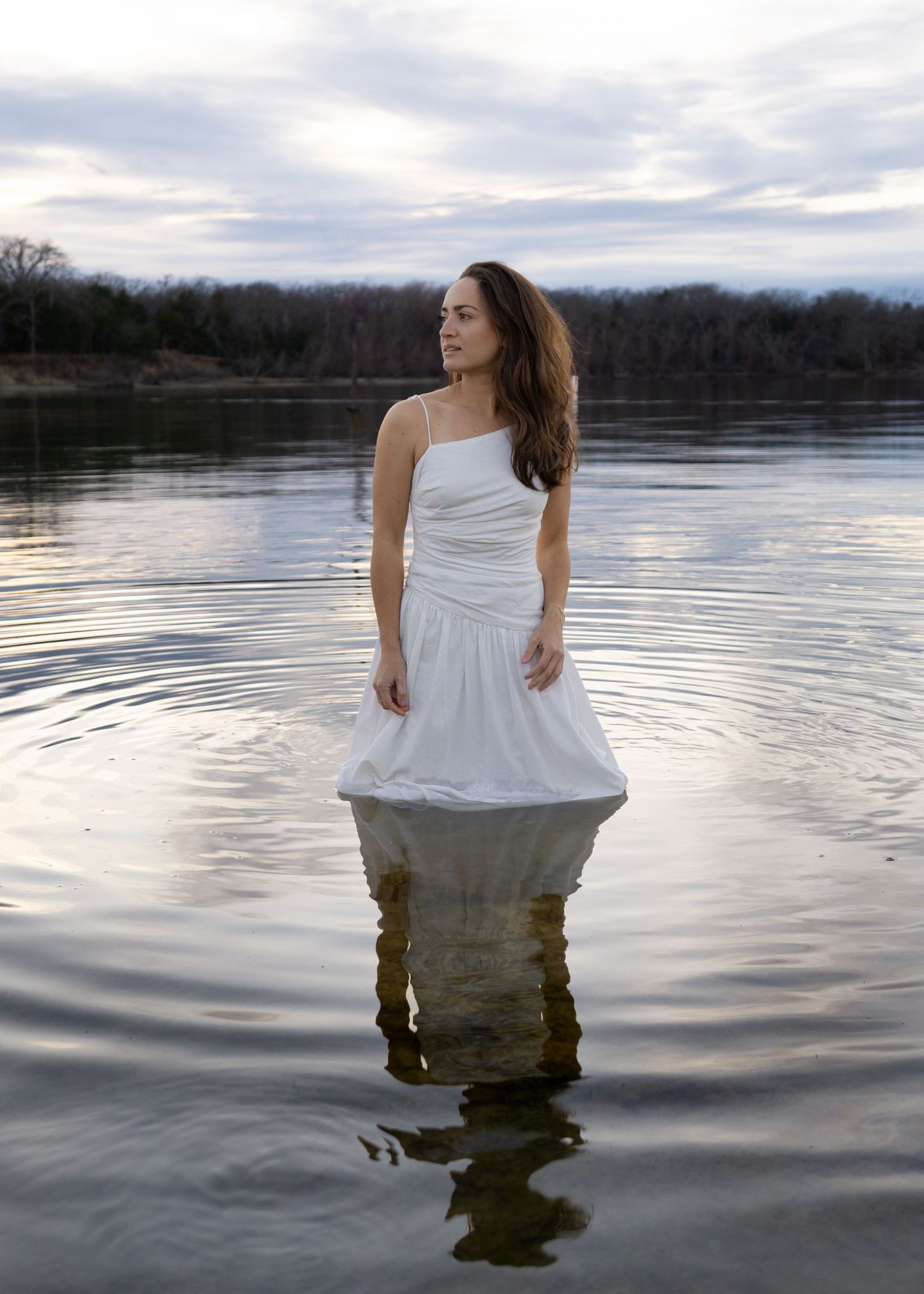 Woman in white dress standing in calm lake water at dusk with trees and cloudy sky reflected on surface.
