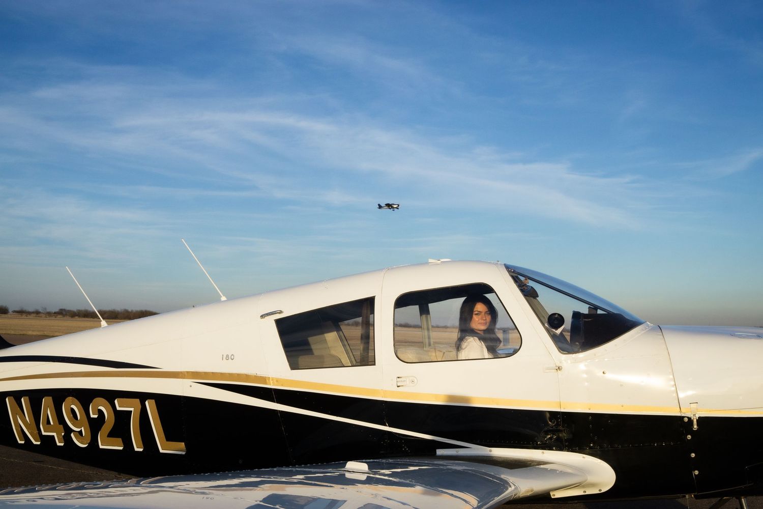 Person sitting in cockpit of small white Piper aircraft N4927L on runway under blue sky.