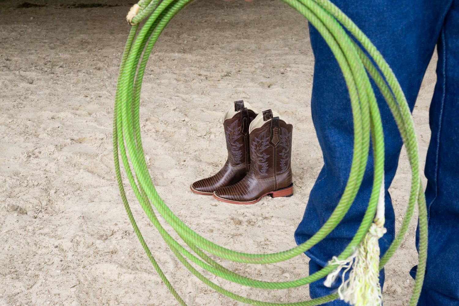 Cowboy boots and green lasso rope held by person in blue jeans on sandy ground.