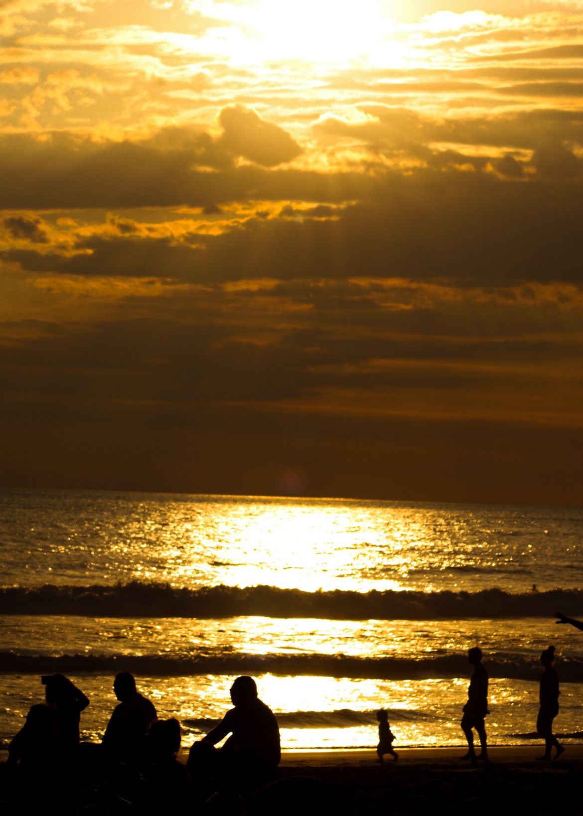 Silhouettes of beachgoers against golden sunset sky and reflective ocean water.