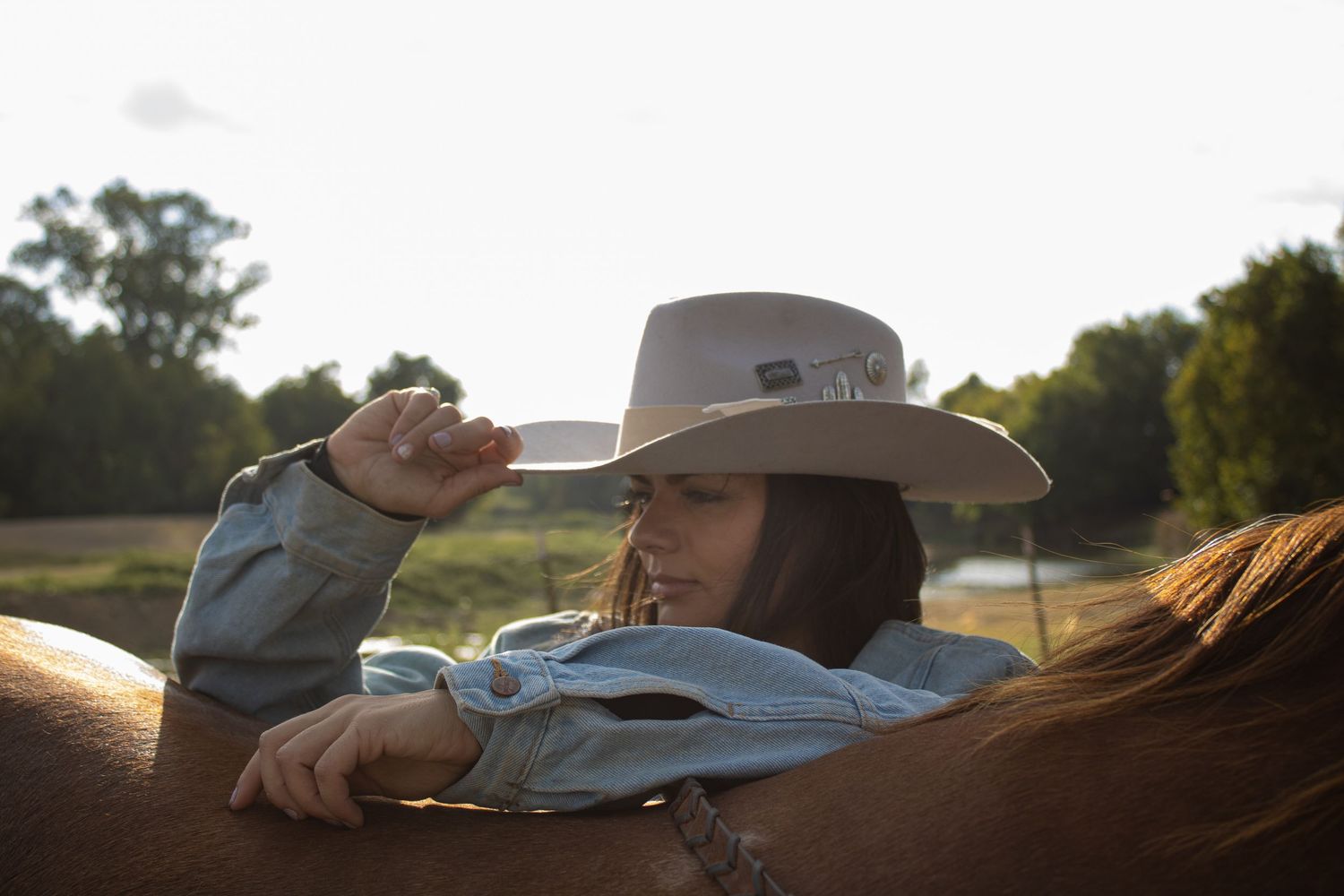 A cowboy in a traditional hat rides horseback across a scenic ranch landscape at sunset.