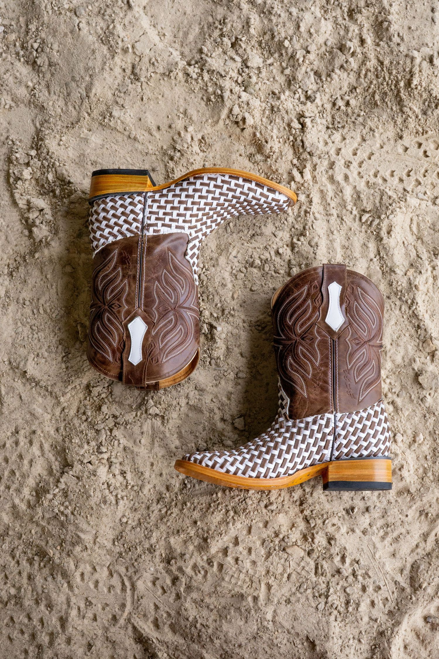 Brown and white woven leather cowboy boots with intricate stitching, resting on sandy ground.