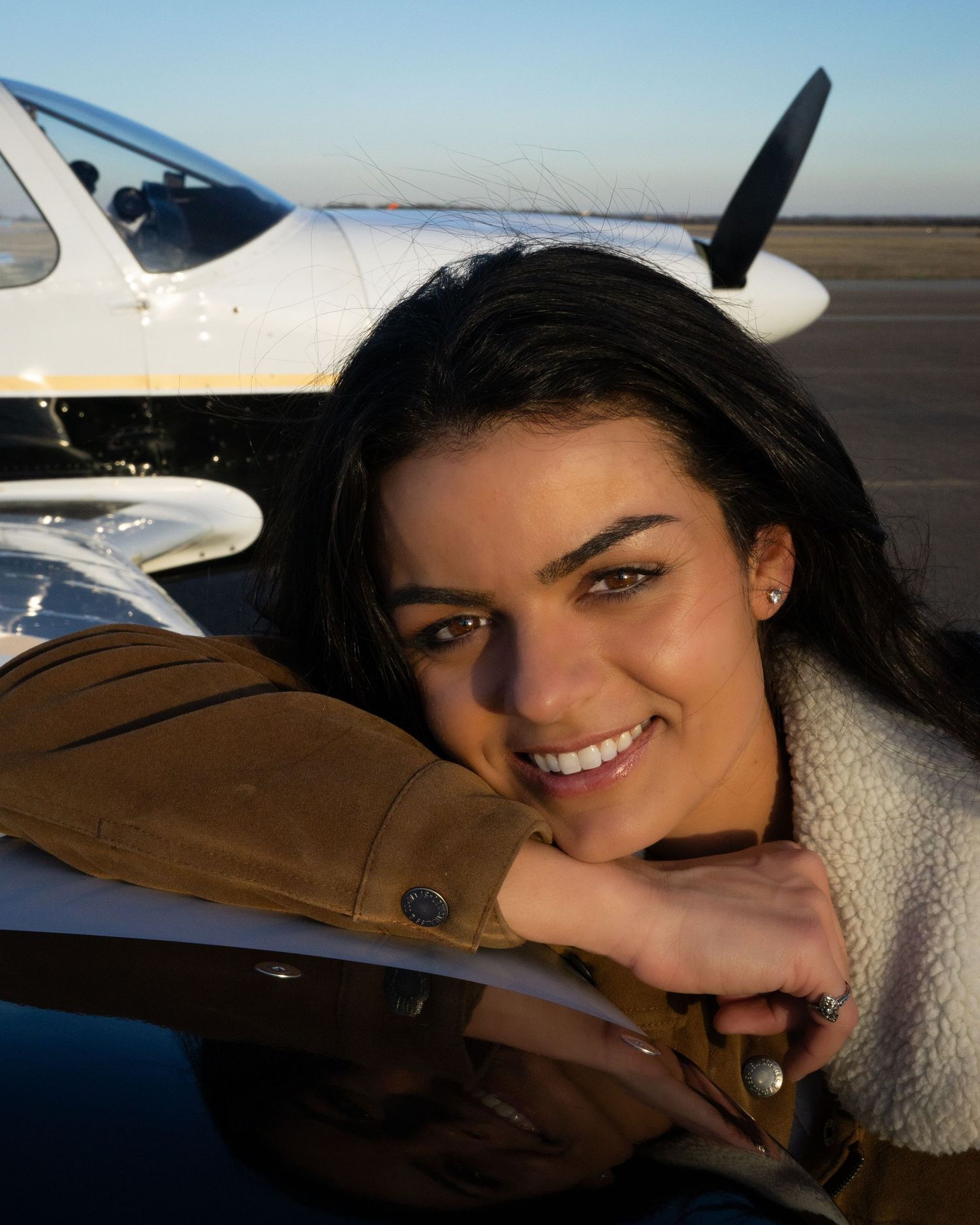 Young woman smiling and leaning on a small white aircraft at an airfield during golden hour.