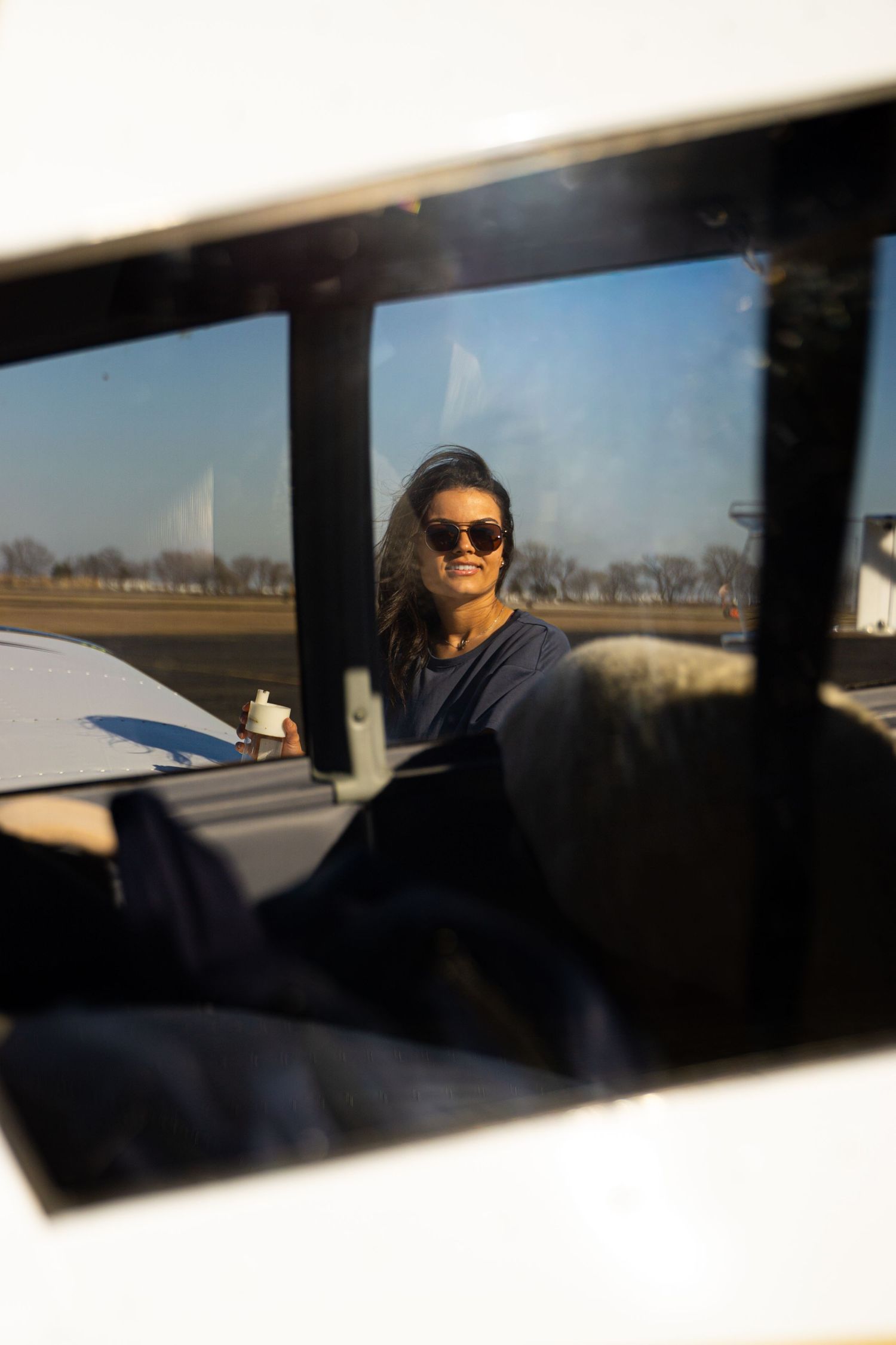 Woman in sunglasses smiling, viewed through a small aircraft window on a sunny day at an airfield.