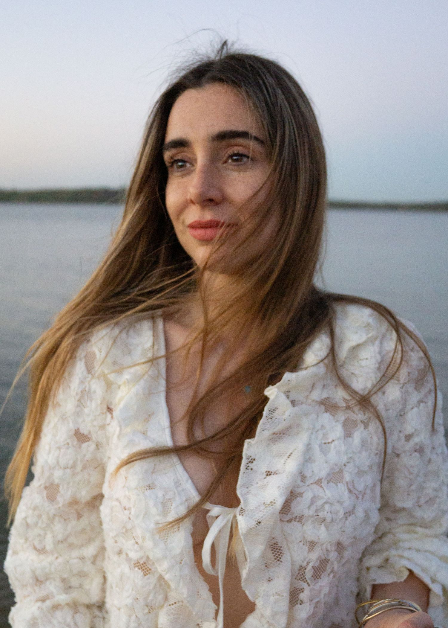 Young woman with long brown hair wearing a white lace jacket poses near a serene waterfront at dusk.