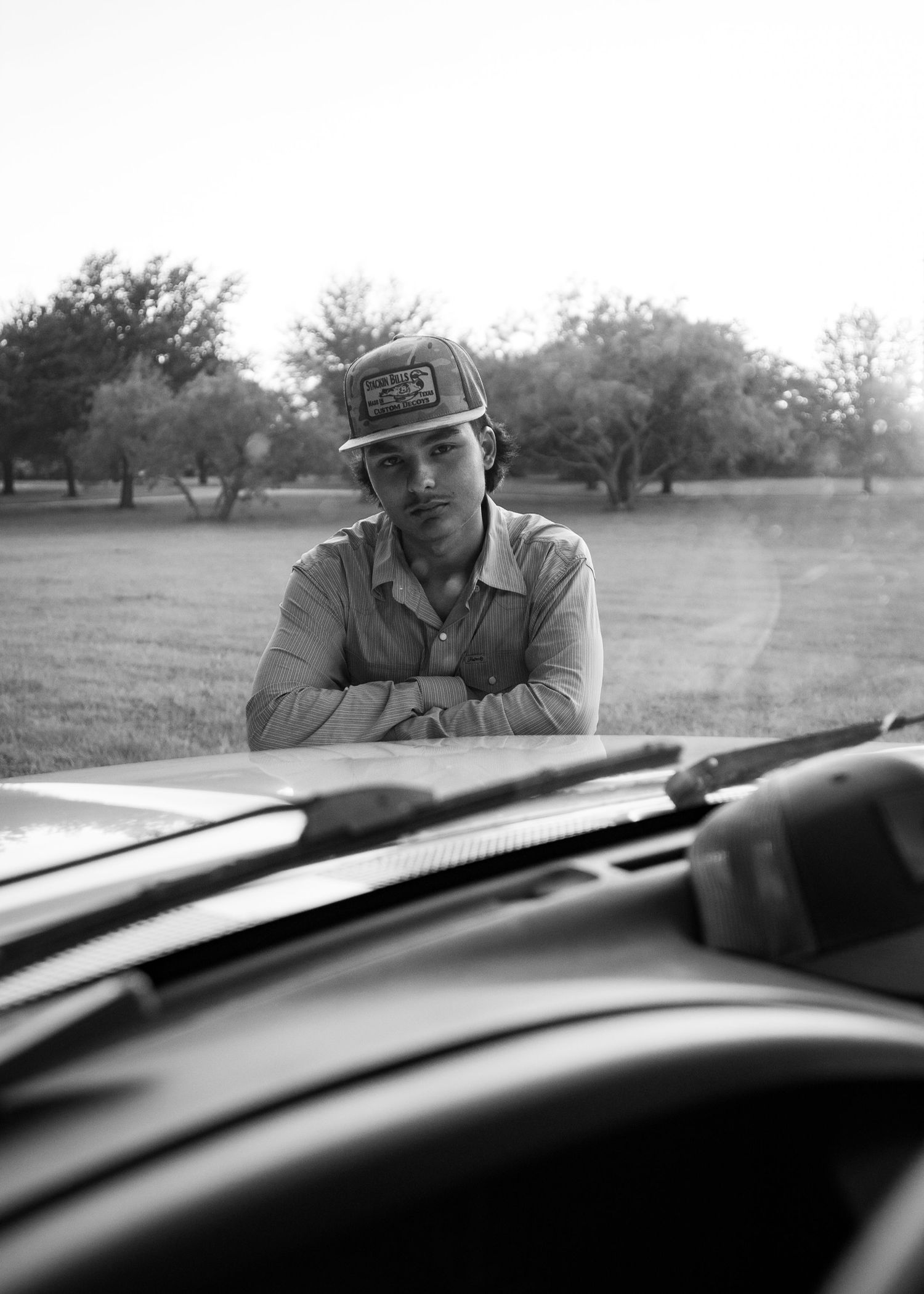 Young man in trucker cap leans on truck hood in black and white rural outdoor photo.