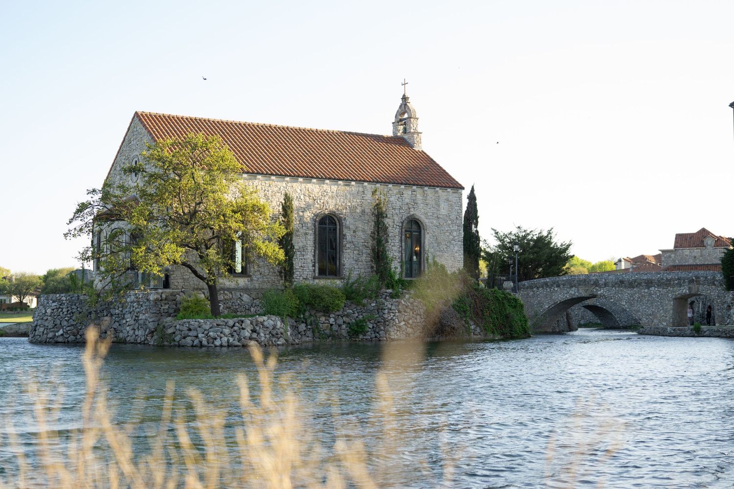 Historic stone chapel on a tranquil island surrounded by water, with a arched bridge and lush trees at golden hour.