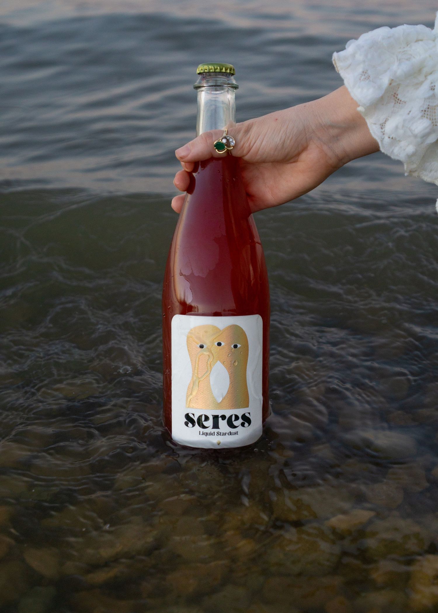Hand holding a bottle of Seres Liquid Sunshine rosé wine over rippling water, wearing a white lace sleeve.