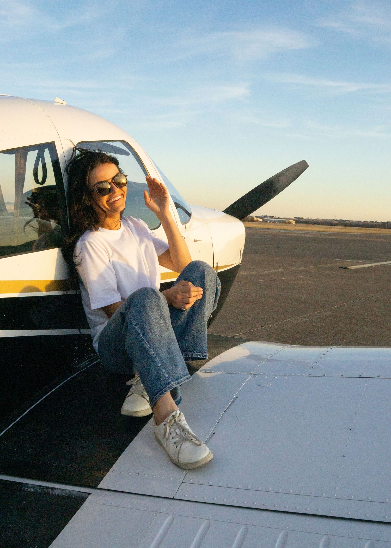 Someone in sunglasses and casual attire sits on a small aircraft wing at an airfield during golden hour.