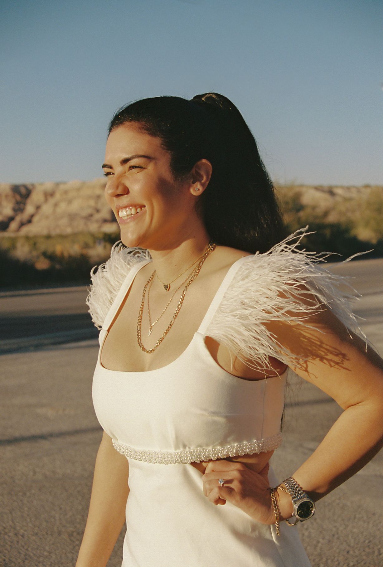 A person in a white dress with feathered sleeves poses against a desert landscape at sunset.