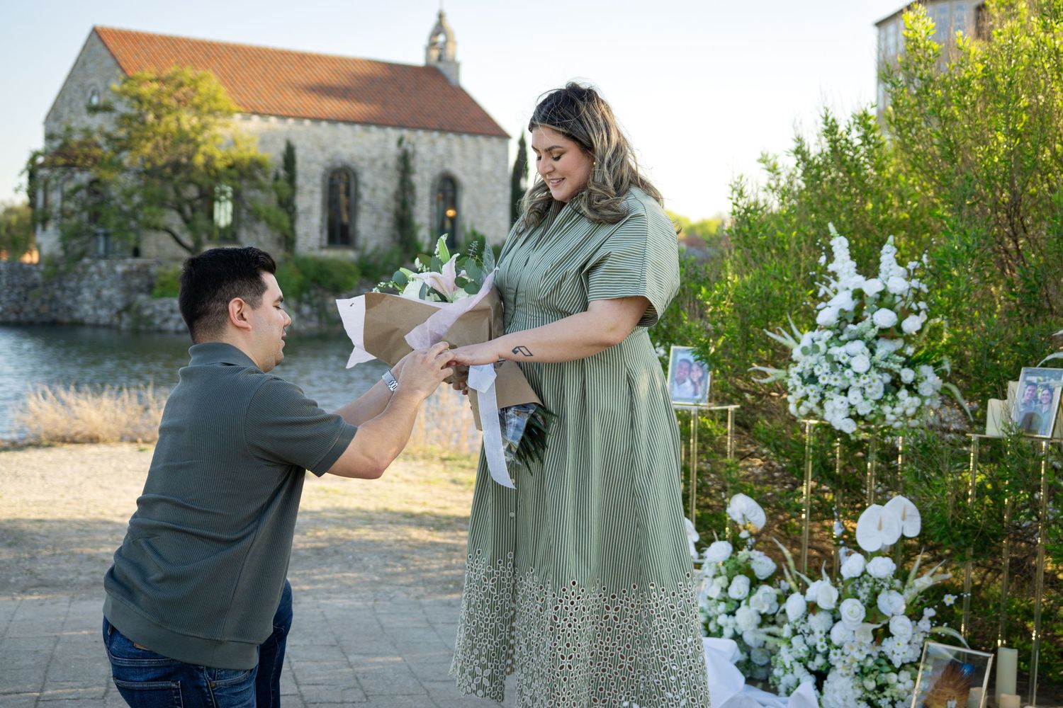 Man kneeling to propose to woman with flowers near a decorated outdoor venue with a church backdrop.