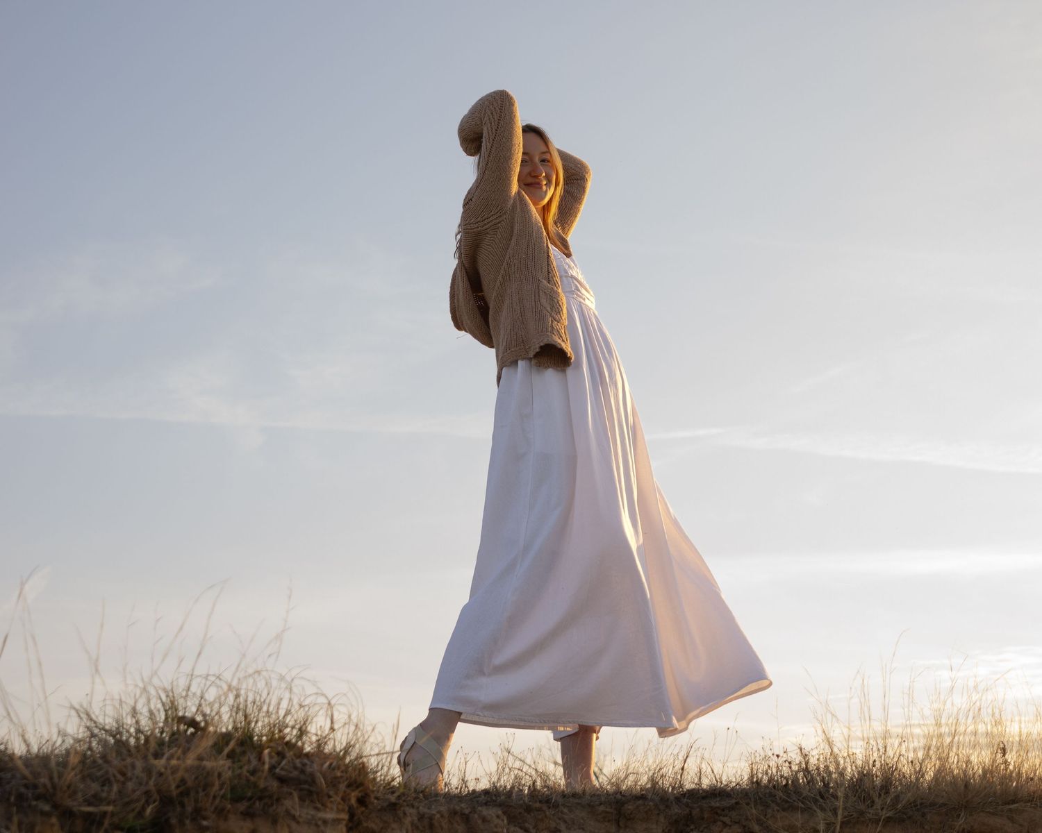 Figure in flowing white dress stands gracefully atop a grassy hill at sunset, creating a dreamy silhouette against the sky.