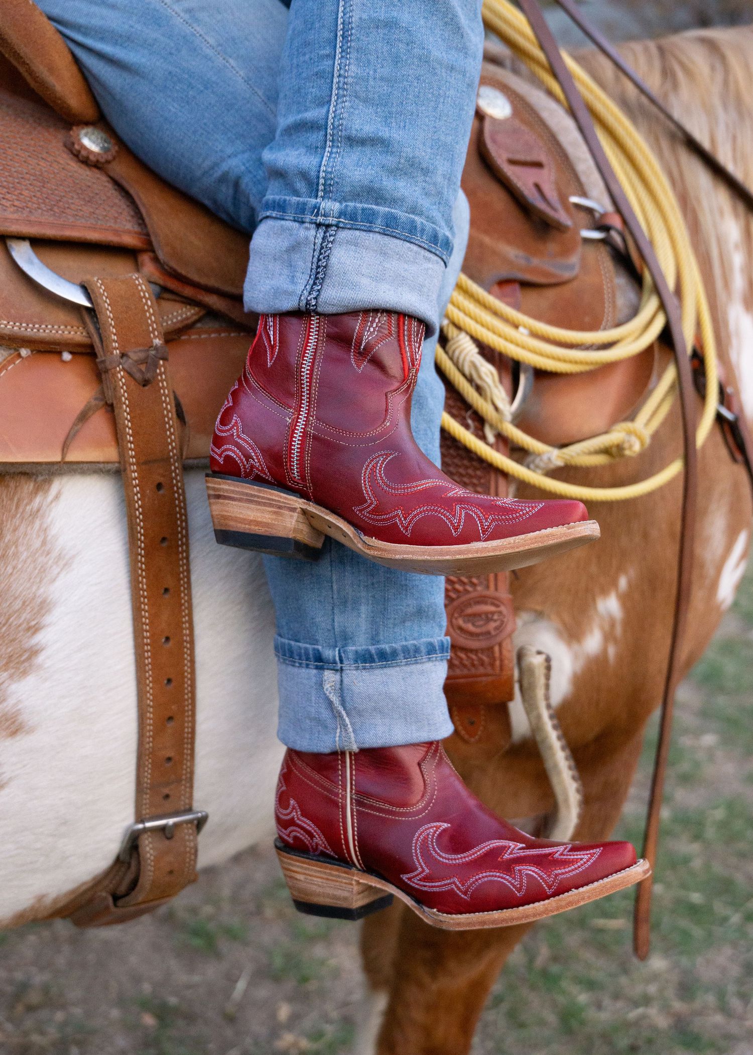 Red leather cowboy boots with decorative stitching shown on horseback with rolled-up jeans and leather saddle details.
