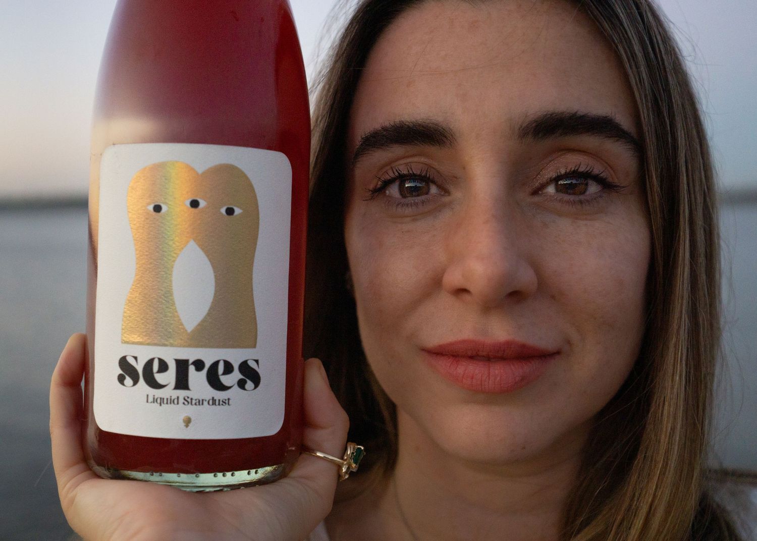Woman holding a bottle of Seres Liquid Stardust pink wine with a whimsical bird label at sunset.