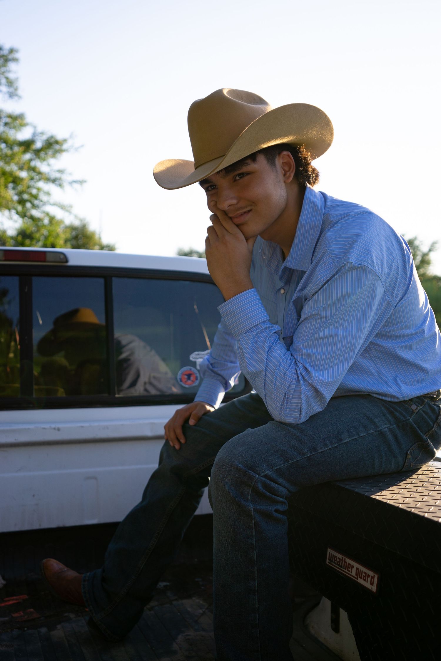 Young man in cowboy hat and jeans sitting on truck tailgate, wearing blue shirt, relaxed pose outdoors.