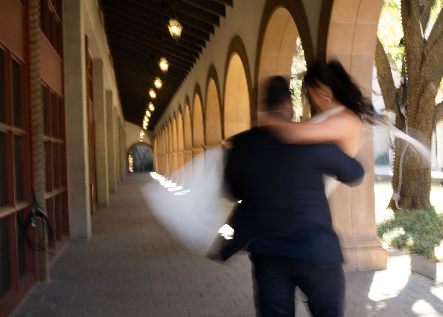 Two people in dark clothing walk down a dimly lit covered walkway with arched columns.