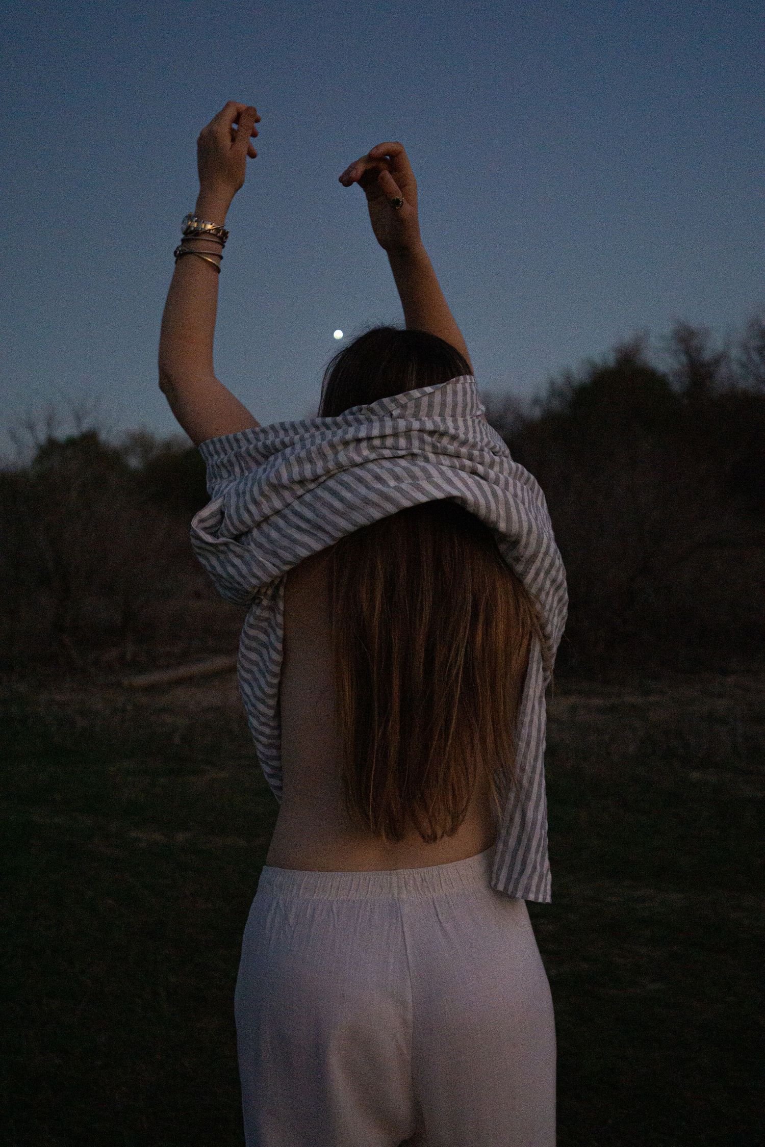 Woman in striped shirt raising arms toward the moon at dusk in a field at twilight.