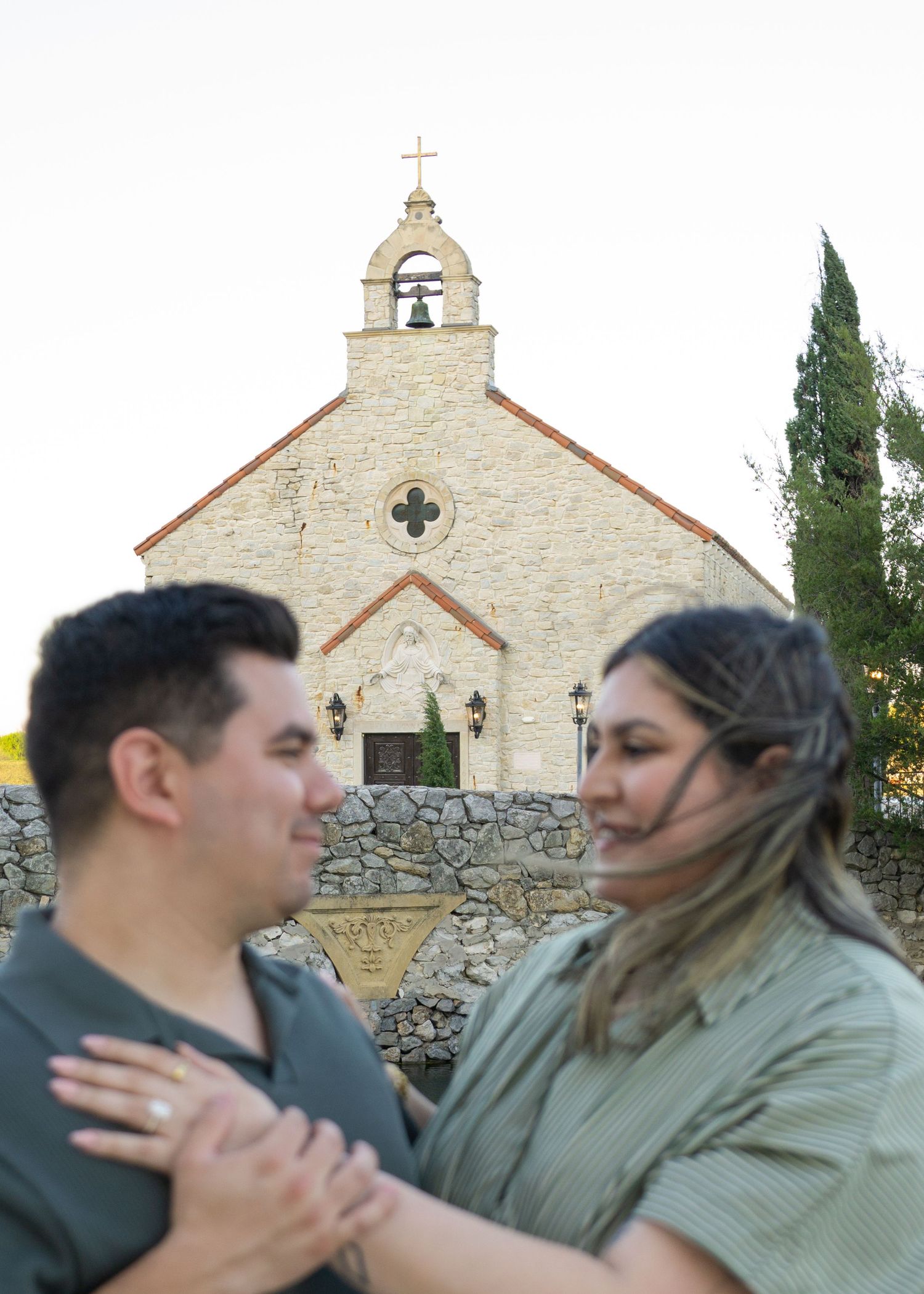 Couple embraces in front of a historic stone chapel with a bell tower and cross during an outdoor engagement photo session.