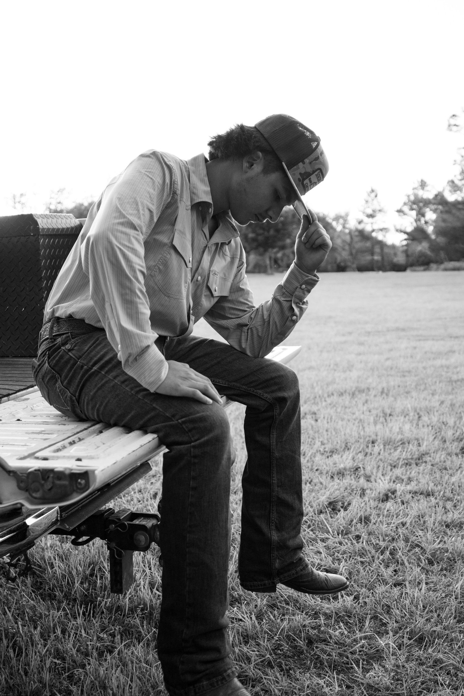 Black and white photo of a man in jeans and cap sitting on a bench, head bowed in contemplation outdoors.