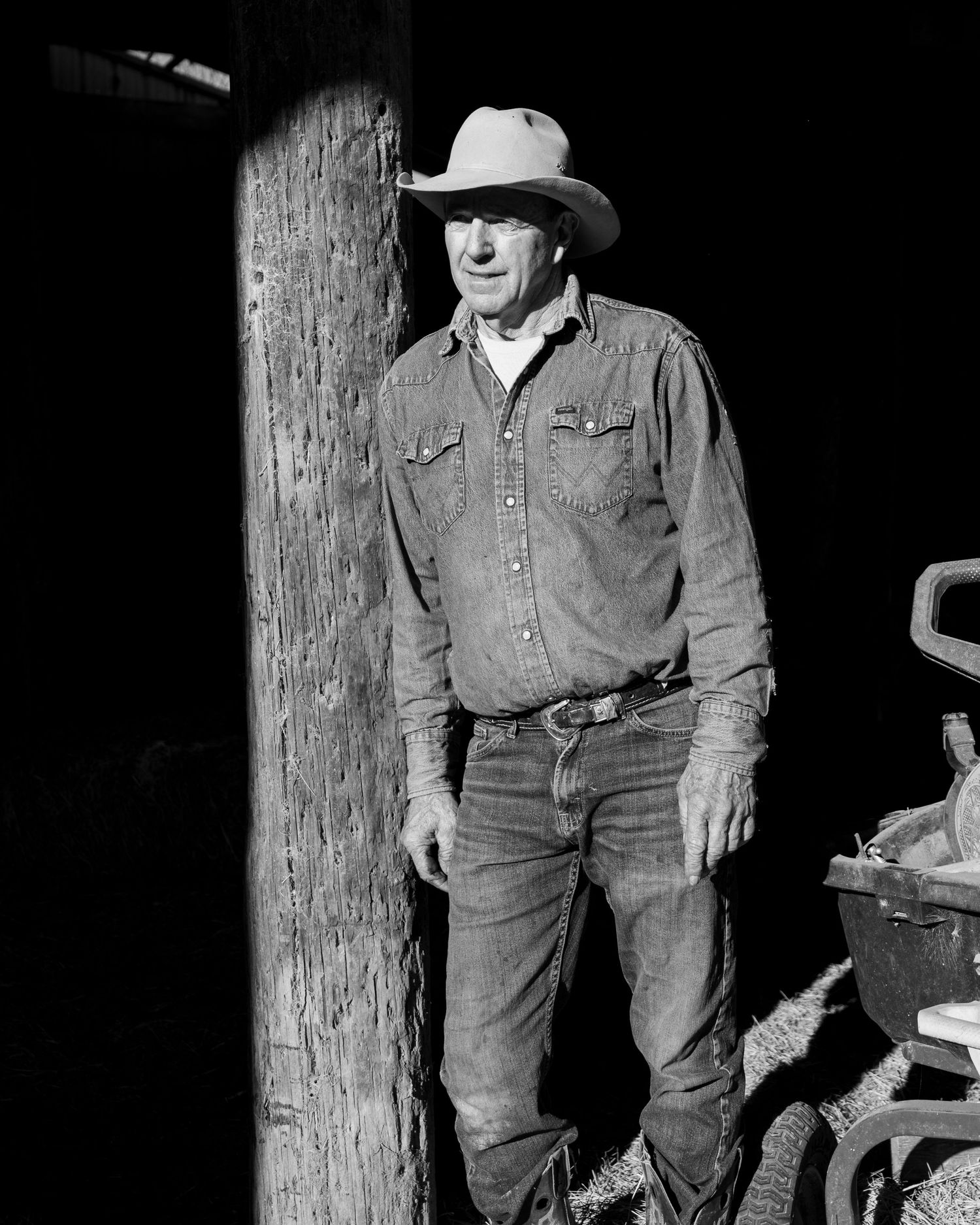 Black and white photo of a rancher in denim and cowboy hat leaning against a wooden post in rustic setting.