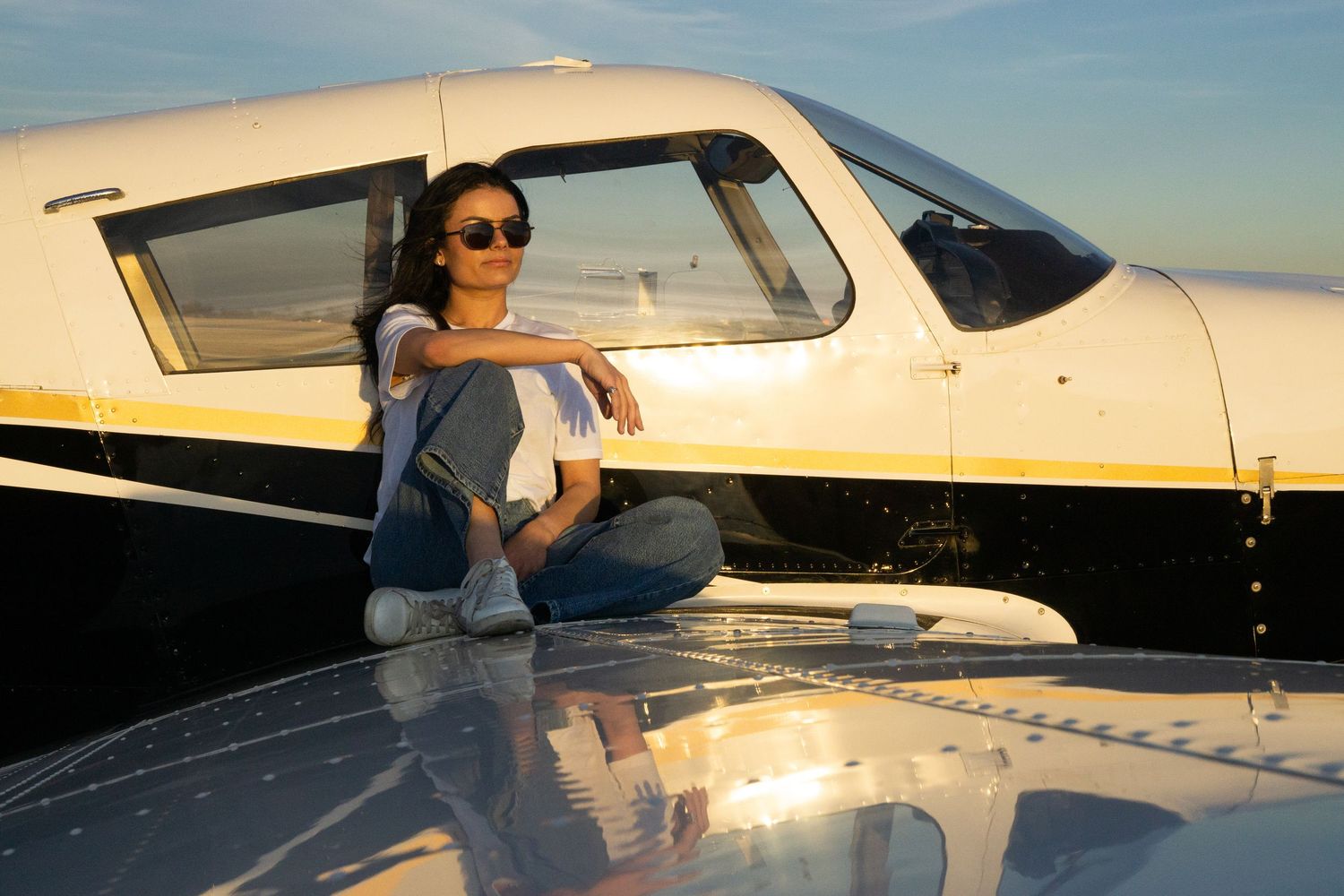 A person in sunglasses sits on the wing of a small black and white private airplane during sunset.