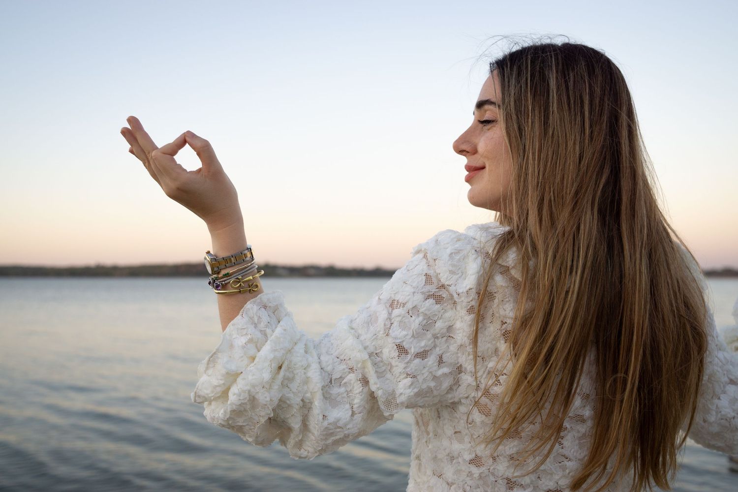 Young woman meditating outdoors by a lake at sunset, making an OK gesture with her hand while wearing a white lace sweater.