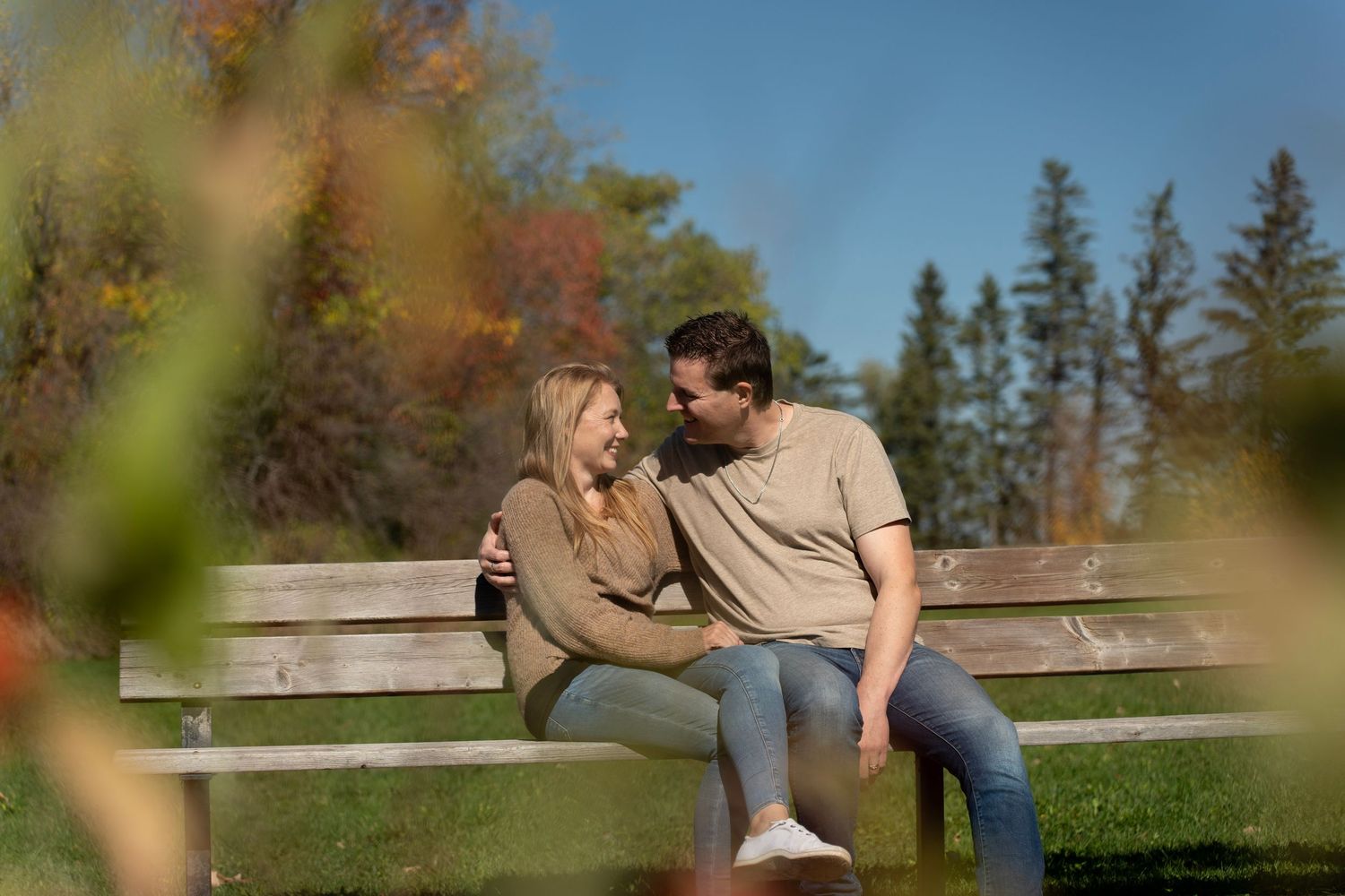 A couple sits close together on a wooden bench in a park during autumn with evergreen trees in the background.